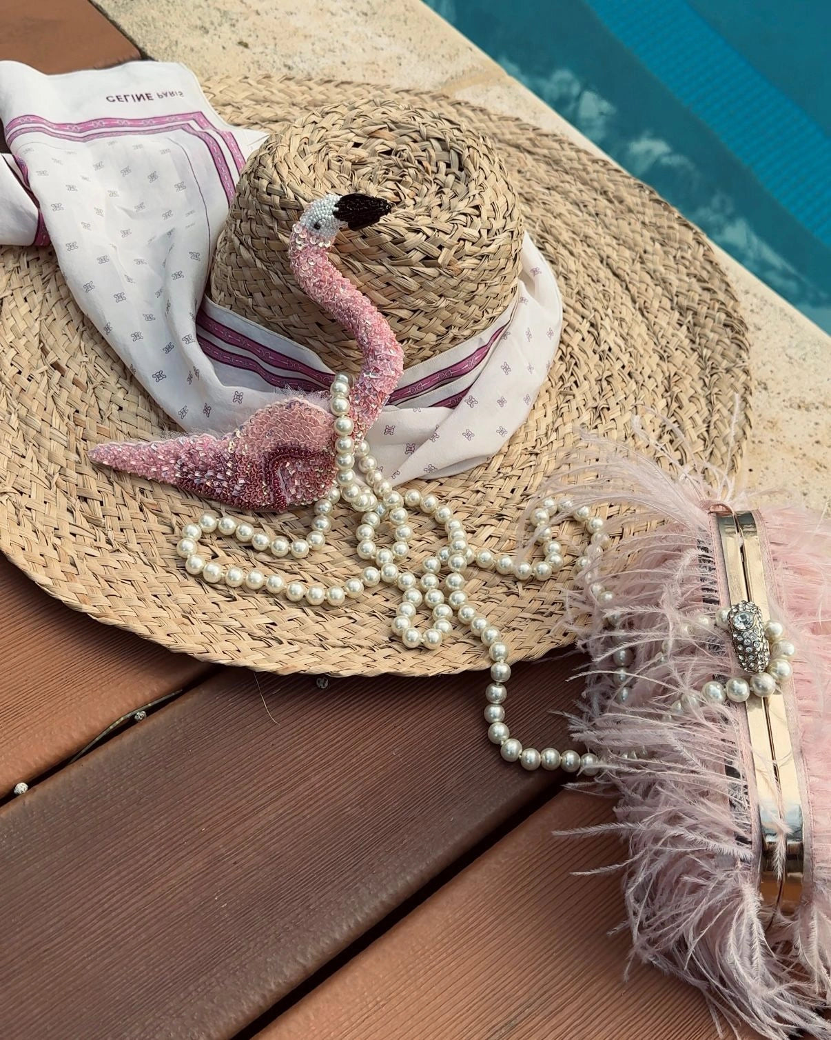 Straw hat with Broochella Tropicana hand beaded pink flamingo brooch, pearls, and feathered clutch on a wooden surface by a pool.