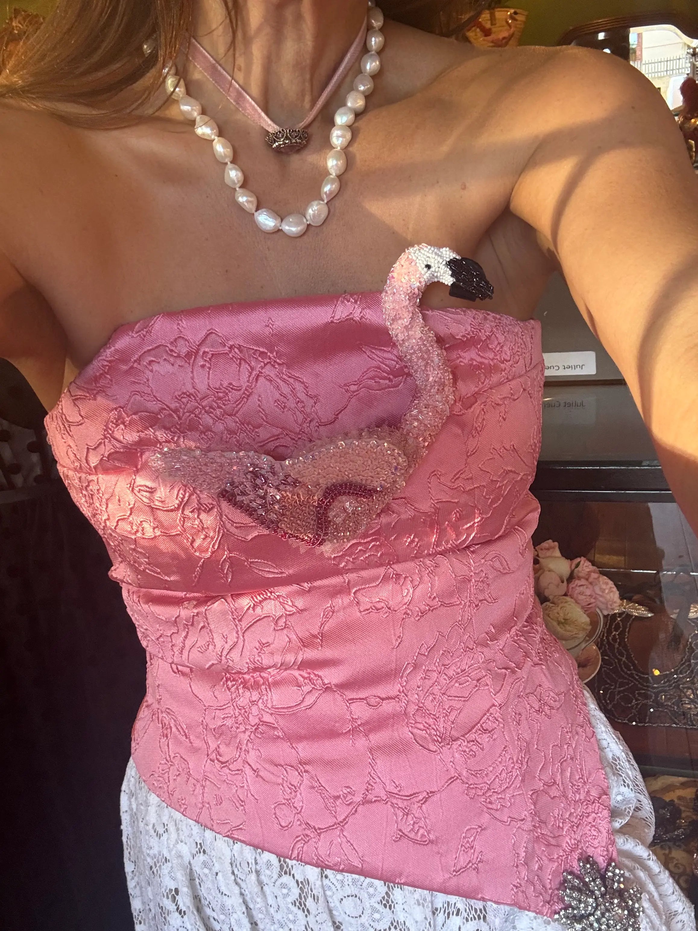 A close-up of a woman wearing a strapless pink satin corset-style top with a large Broochella tropicana pink flamingo brooch on the front, paired with a white lace skirt and layered pearl necklaces.