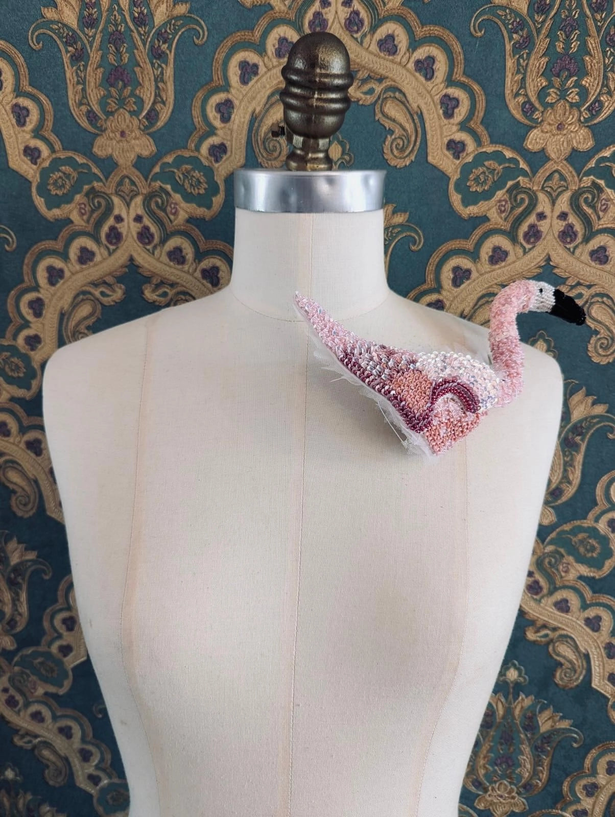 Broochella Tropicana hand beaded 3D figural flamingo brooch. It is pinned to a mannequin. The background is green and gold damask wallpaper.