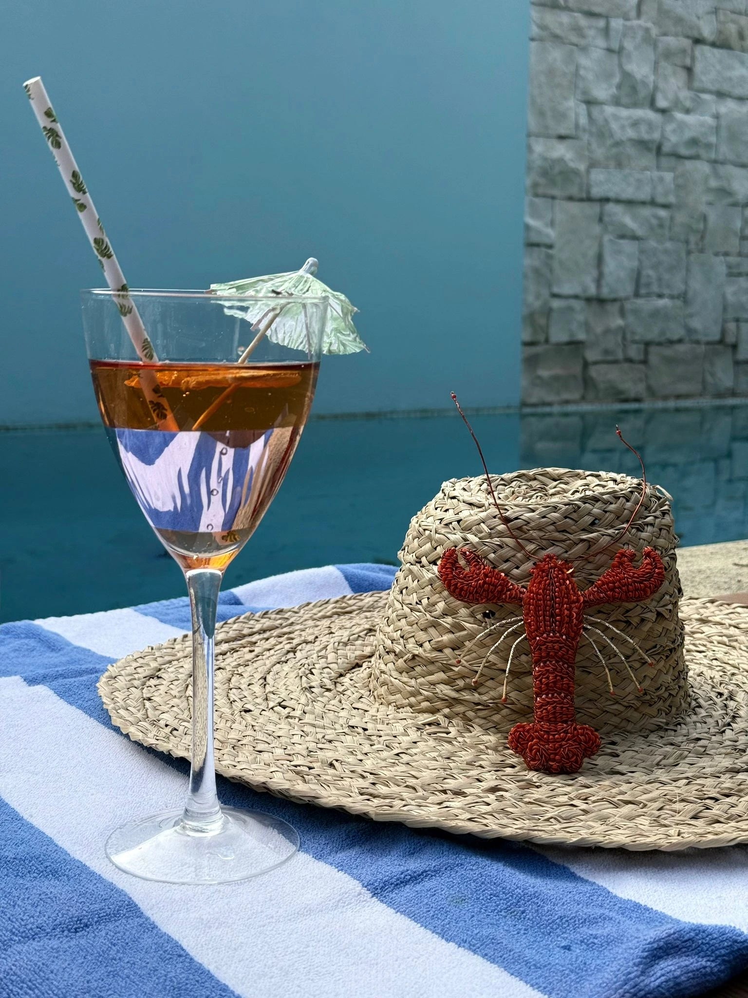 Broochella Tropicana hand beaded red lobster brooch. It is pinned to a straw hat, which is placed on a blue and white striped towel next to a pool. Next to the hat is a cocktail with a straw and an umbrella