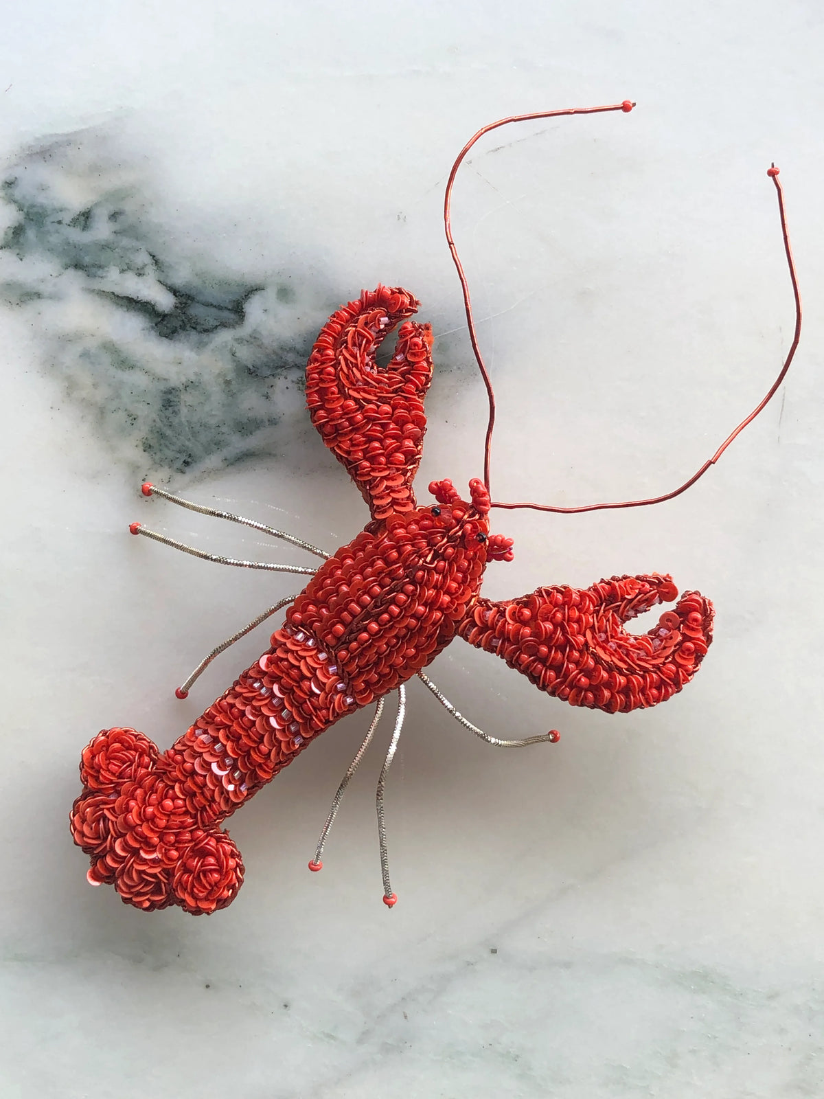 Broochella Tropicana Lobster Brooch Pin, an intricately hand-beaded red lobster statement brooch with sequins, sculptural claws and long antennae, photographed on white marble with soft grey-green veining.