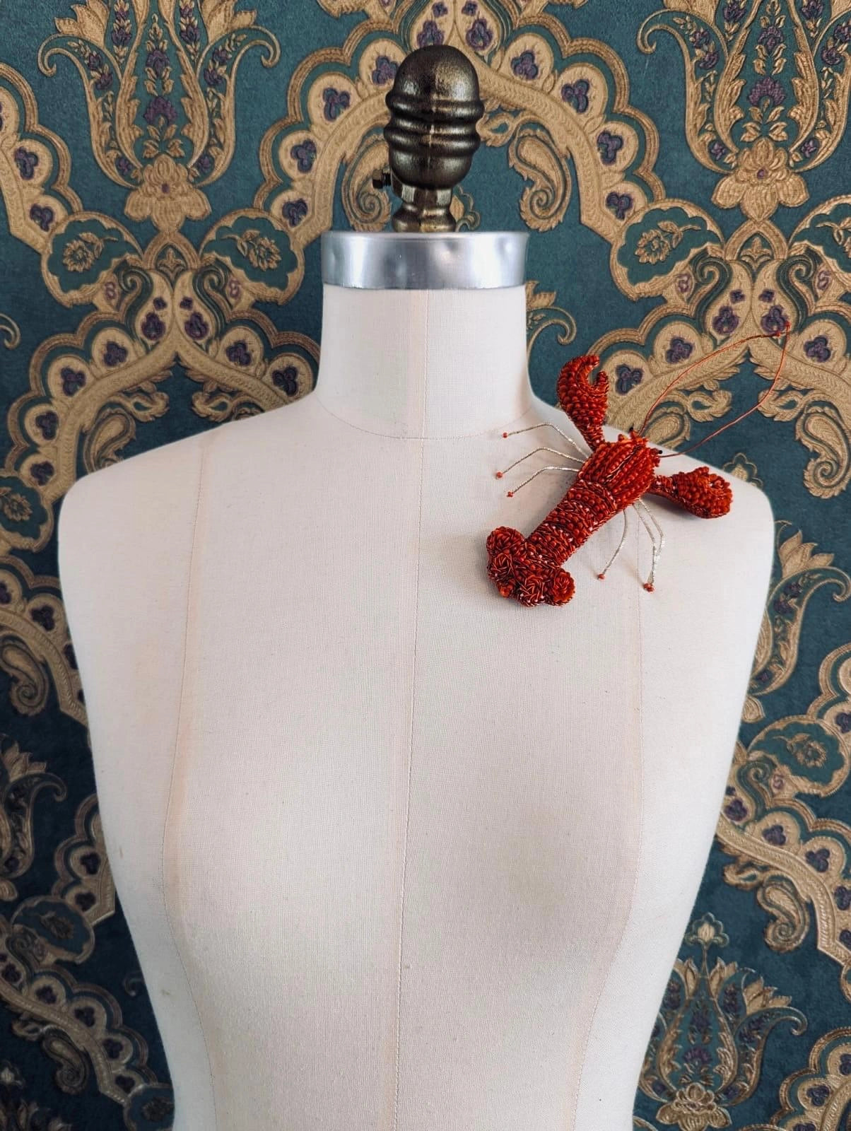 Broochella Tropicana hand beaded 3D figural lobster brooch. It is pinned to a mannequin. The background is green and gold damask wallpaper.