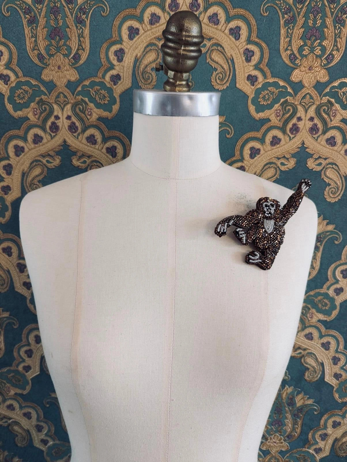 A Broochella Tropicana hand beaded  brown monkey-shaped brooch, displayed on the left shoulder of a cream-colored mannequin against a teal and gold patterned wallpaper background.