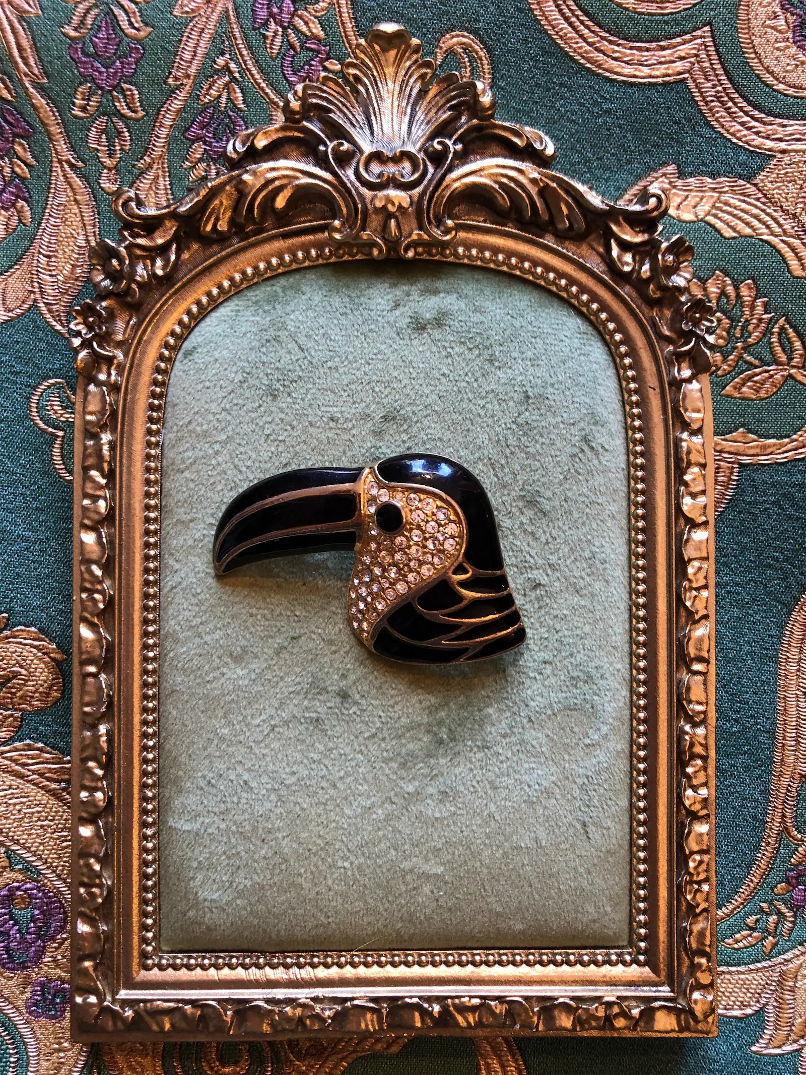 A Broochella Toucano Nero bird-shaped brooch with a glossy black beak and body, a gold-toned patterned head, and small clear rhinestones, displayed inside an ornate antique-style gold frame against a patterned green fabric background.