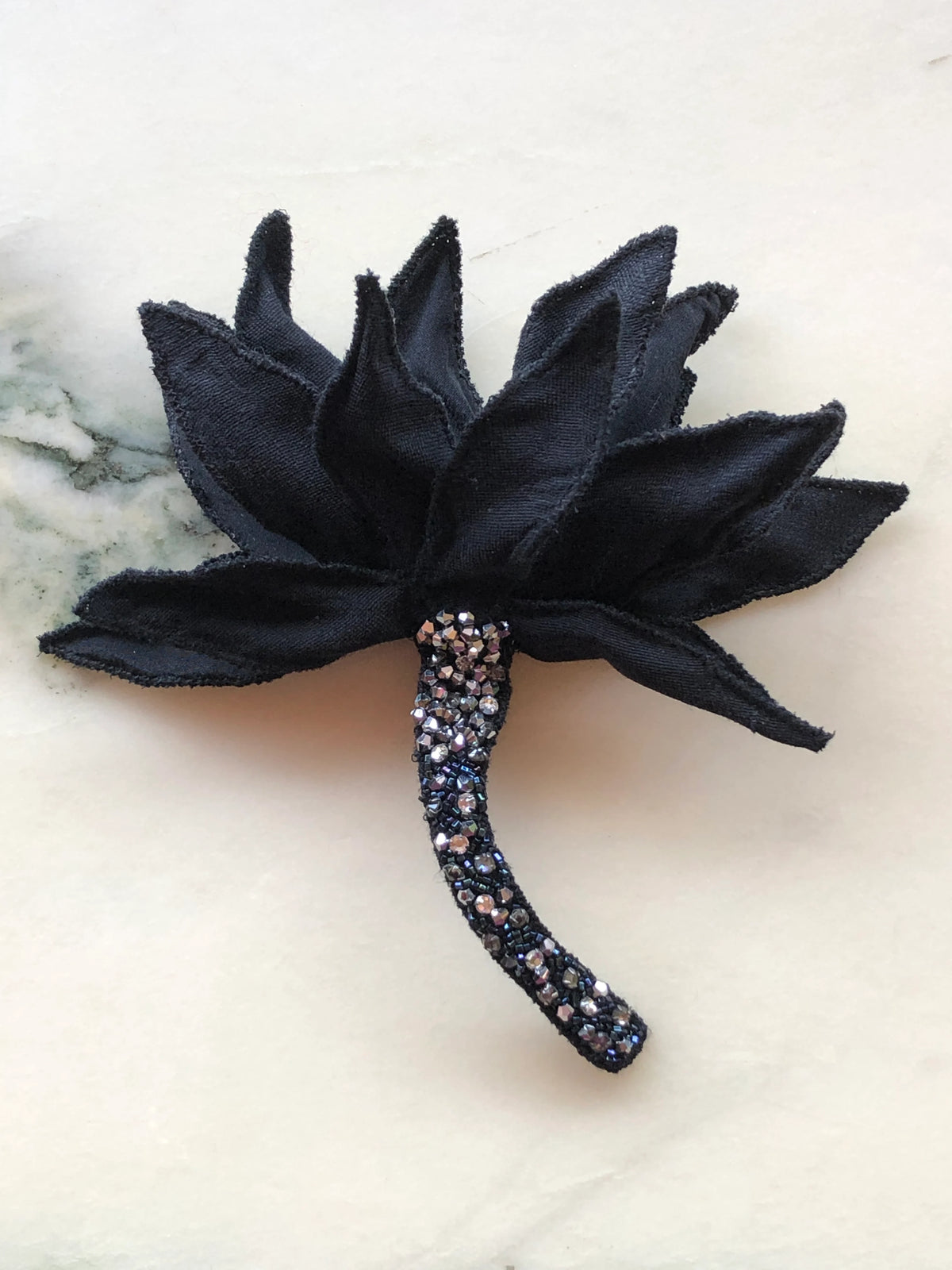 Broochella Tulipano Ingioiellato large black flower brooch pin, featuring dramatic sculptural pointed fabric petals and a long curved stem embellished with black beads and sparkling crystals, photographed on a white marble background with soft grey-green veining.