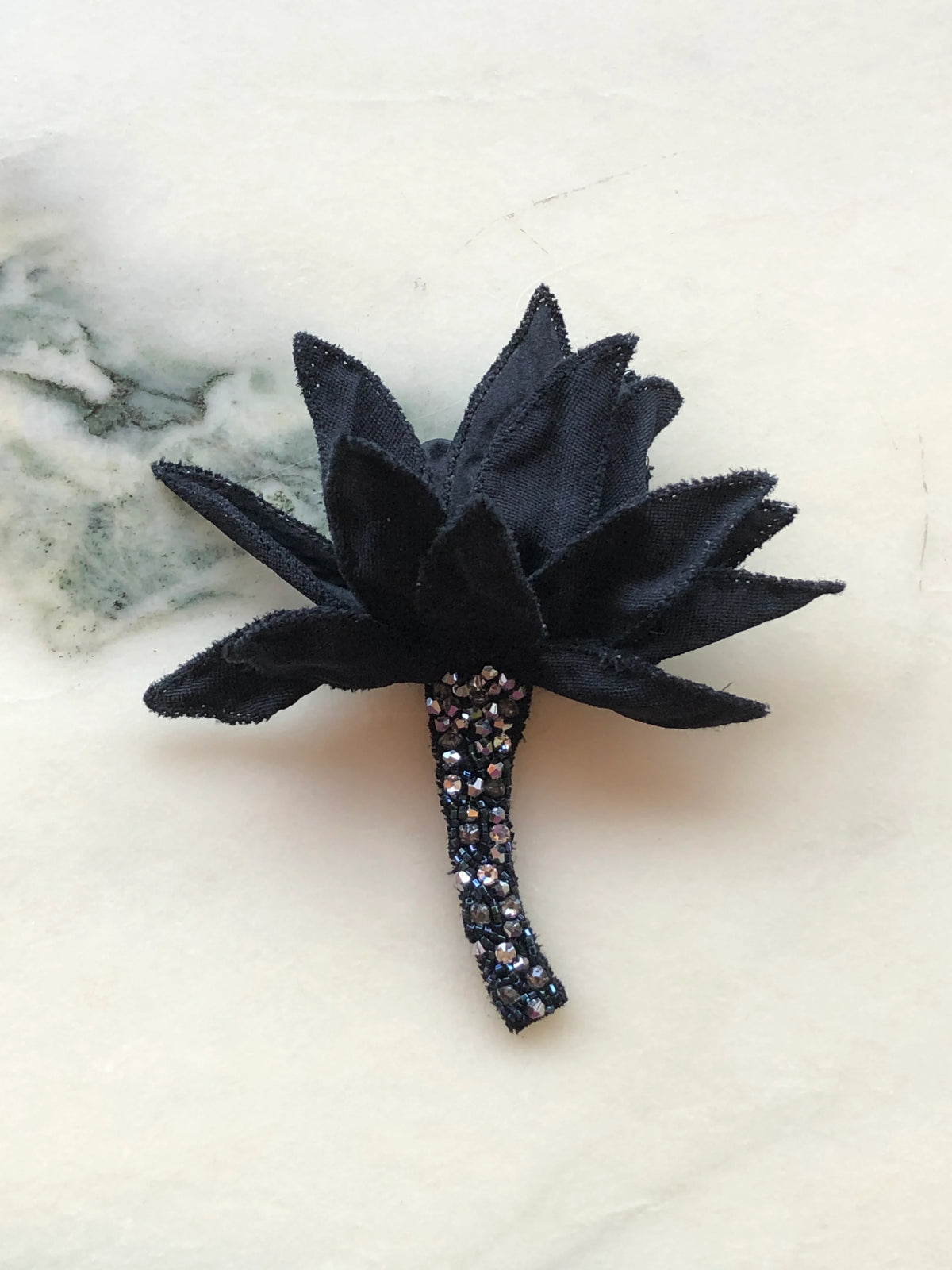 Broochella Tulipano Ingioiellato small black flower brooch pin, featuring sculptural pointed fabric petals and a curved stem embellished with black beads and sparkling crystals, photographed on a white marble background with soft grey-green veining.