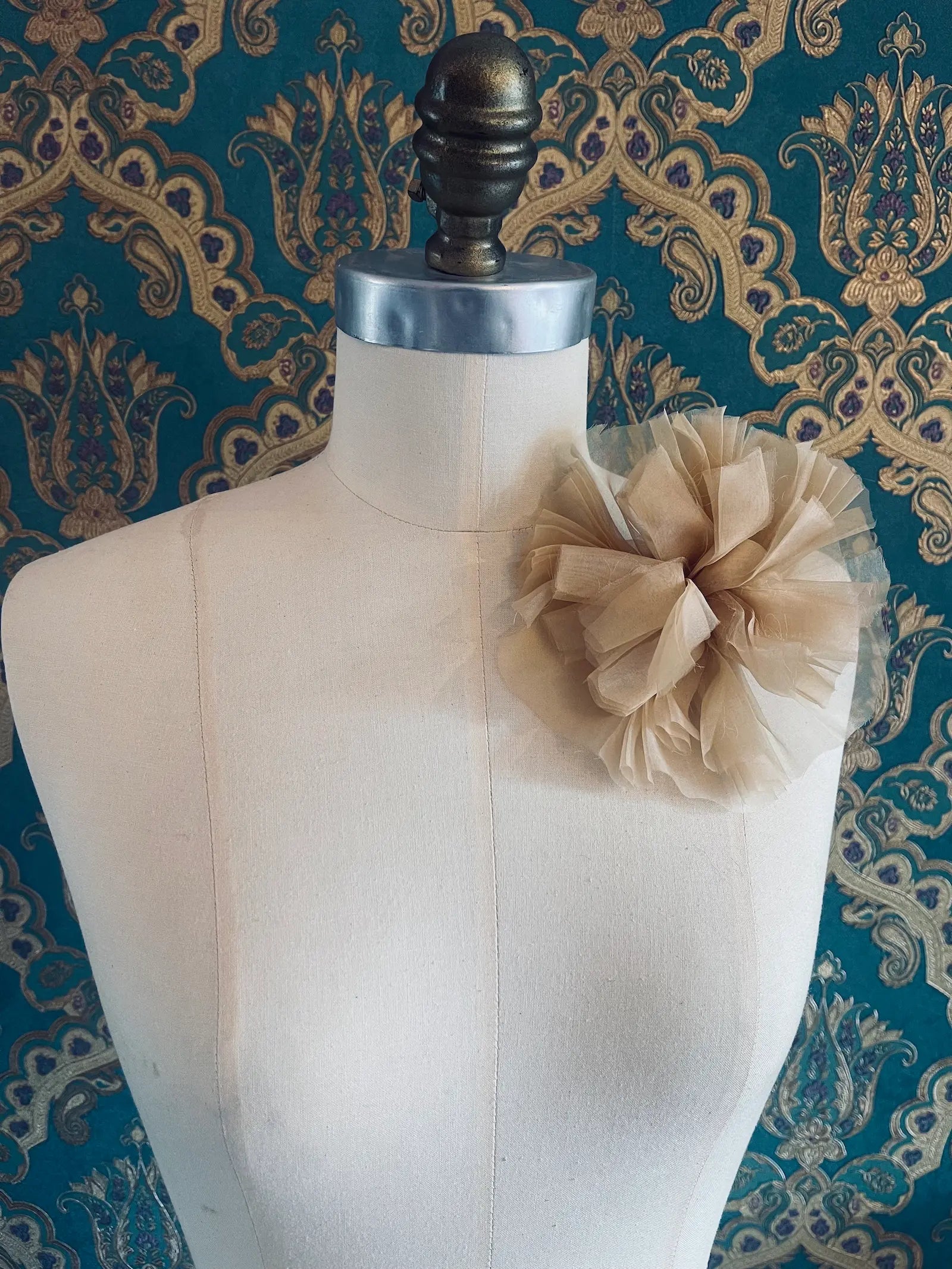Broochella beige handmade fabric flower brooch. Pictured on a mannequin. The background is green and gold damask wallpaper