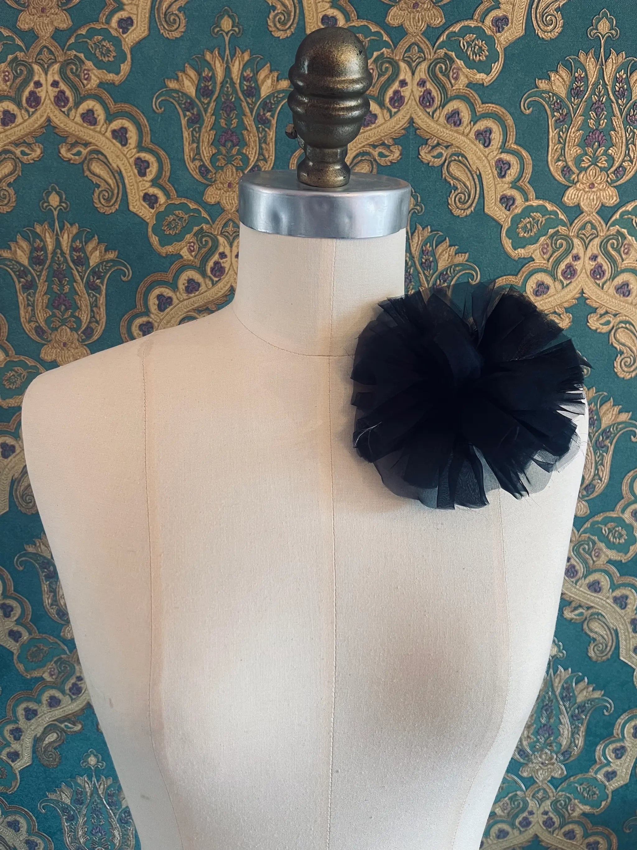 Broochella black handmade fabric flower brooch. Pictured on a mannequin. The background is green and gold damask wallpaper