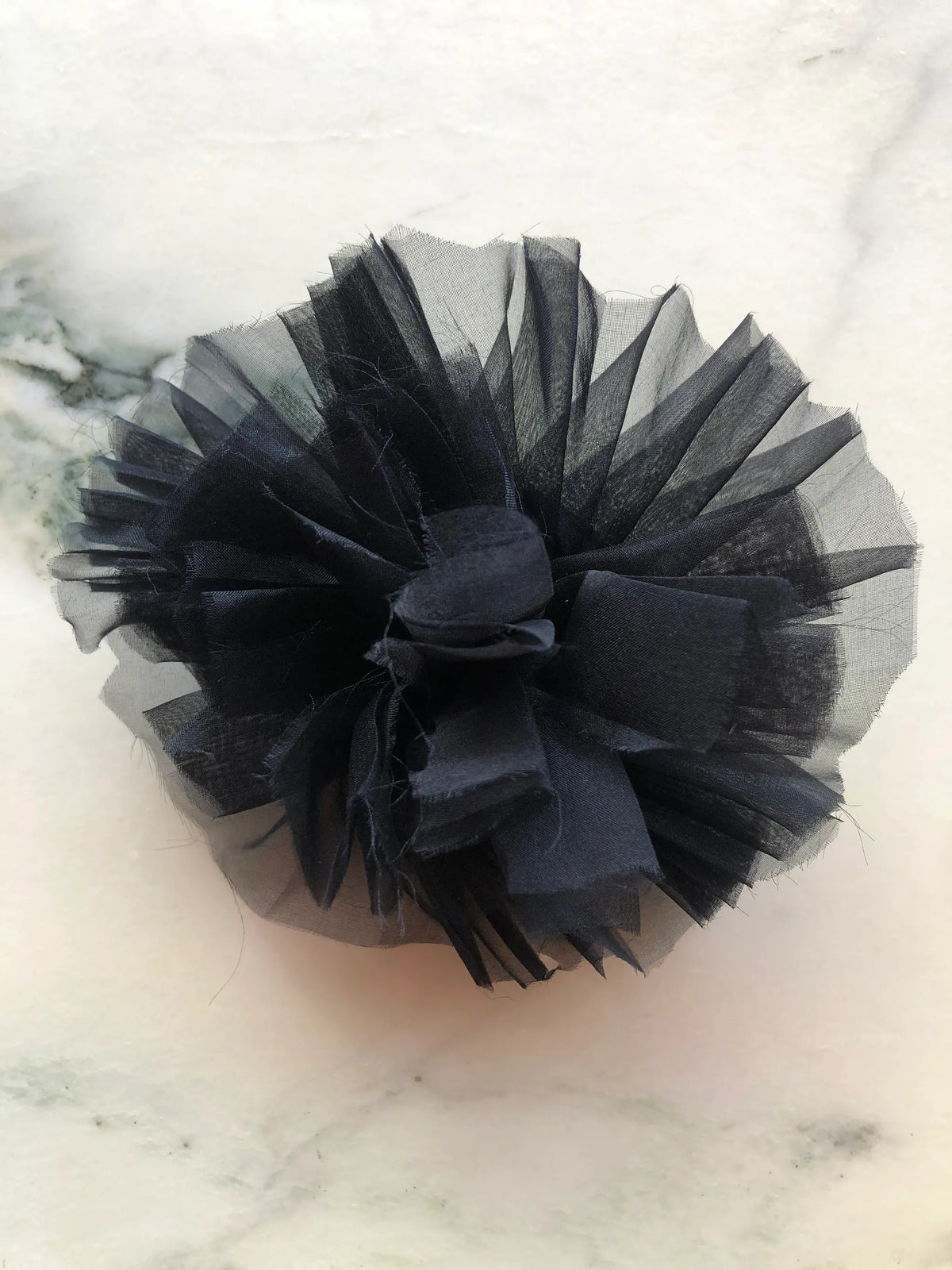 Broochella Tulle Flower Brooch Pin in black, featuring layers of pleated sheer tulle with a sculptural fabric centre, photographed on a white marble background with soft grey-green veining.