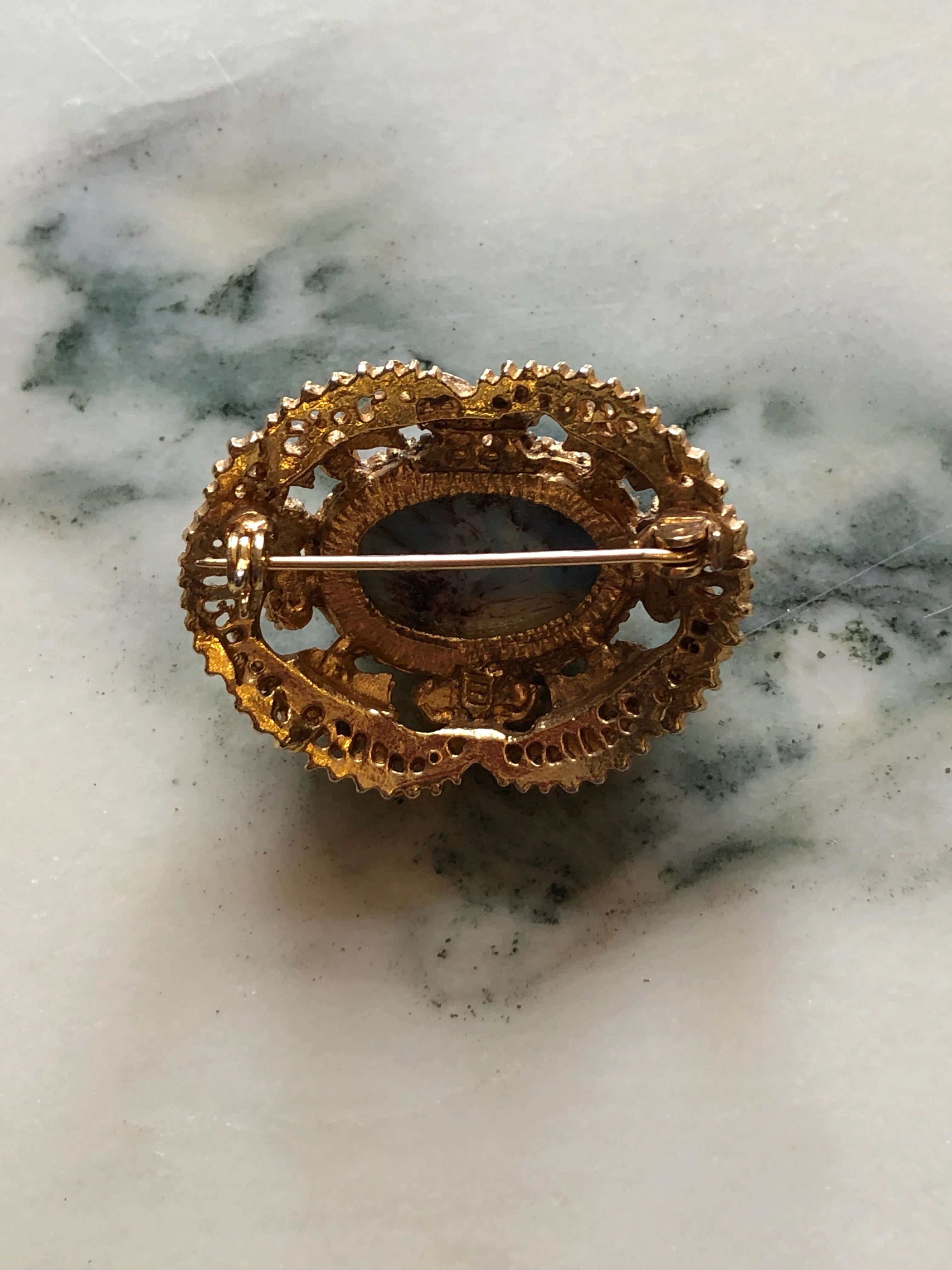 The back view of Broochella Turchese vintage gold-tone brooch with a turquoise-blue marbled cabochon, ornate filigree and faux pearl accents The background is white marble