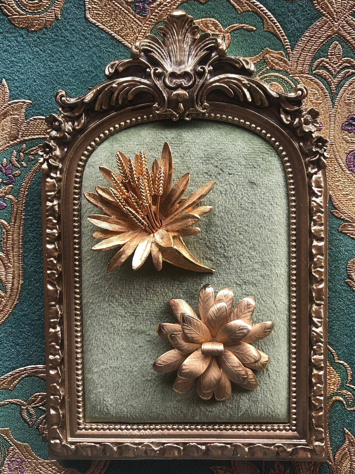 Broochella Two Golden Blooms Vintage Brooch Set featuring two sculptural gold-tone floral brooches displayed on sage green velvet in an ornate antique gold frame against green and gold patterned fabric.