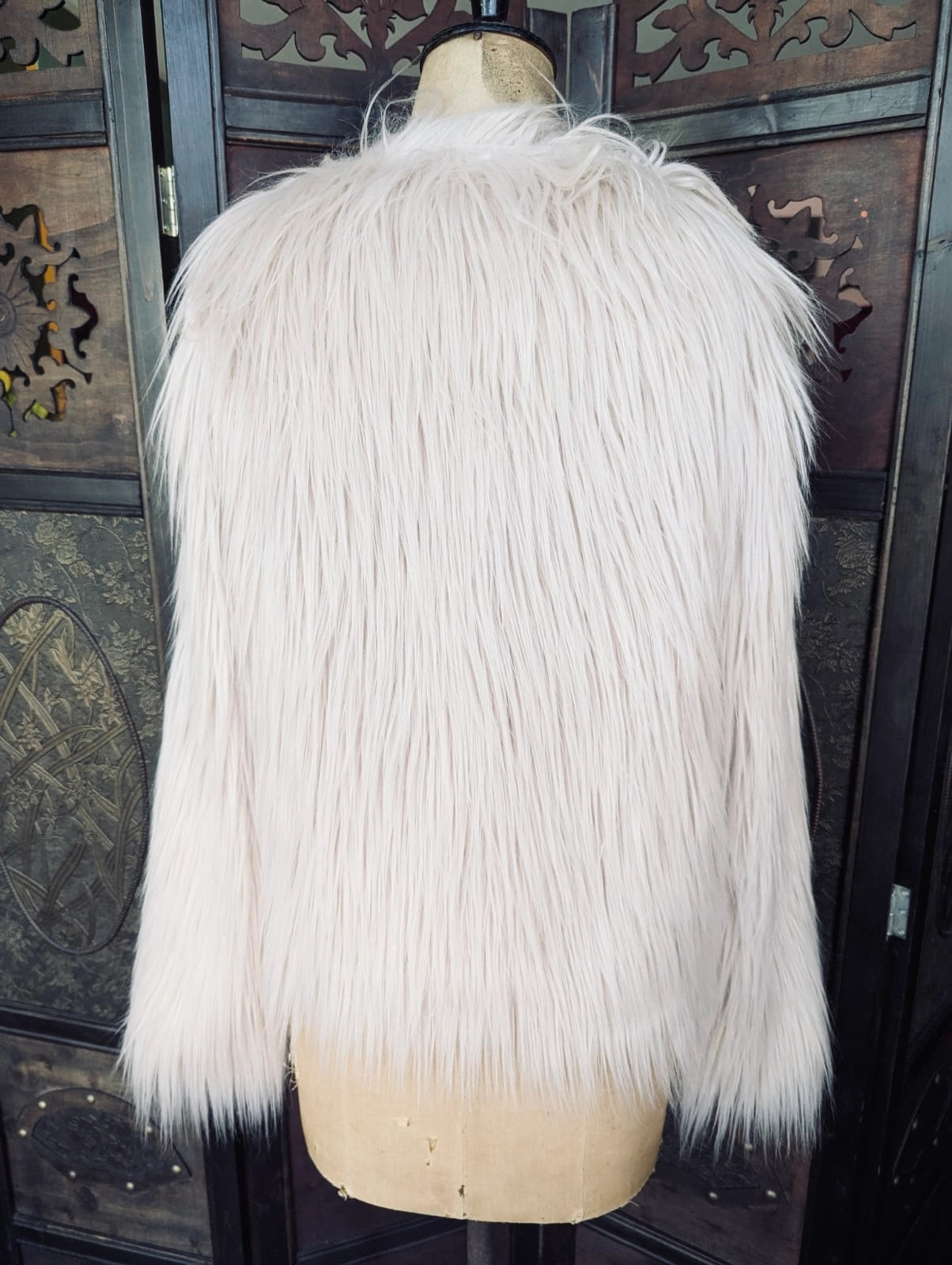 Broochella Unreal Fur Faux fur coat in beige. It is pictured on a vintage mannequin. The background is an ornate, dark wood room divider. 