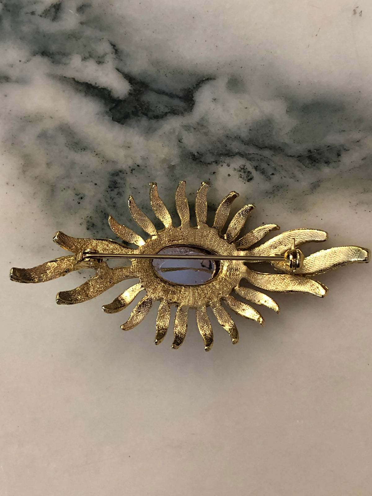 The back view of Broochella Valentina Vintage Purple Sunburst Brooch featuring a deep-purple oval cabochon surrounded by gold-tone crystal-set rays on a cream and grey marble background.