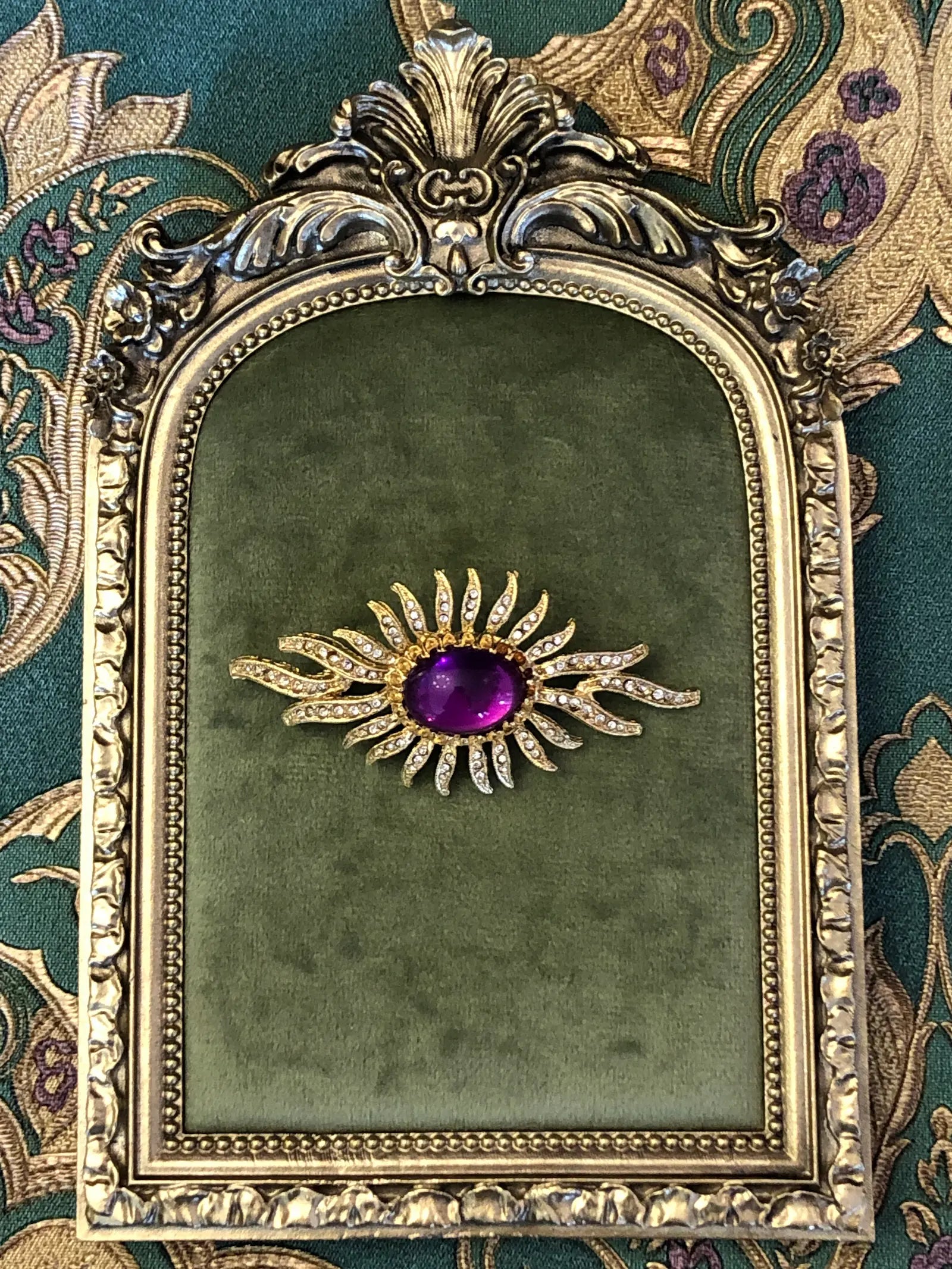 Broochella Valentina Vintage Purple Sunburst Brooch featuring a deep-purple oval cabochon surrounded by gold-tone crystal-set rays. It is displayed on a green velvet tray with an ornate gold frame.