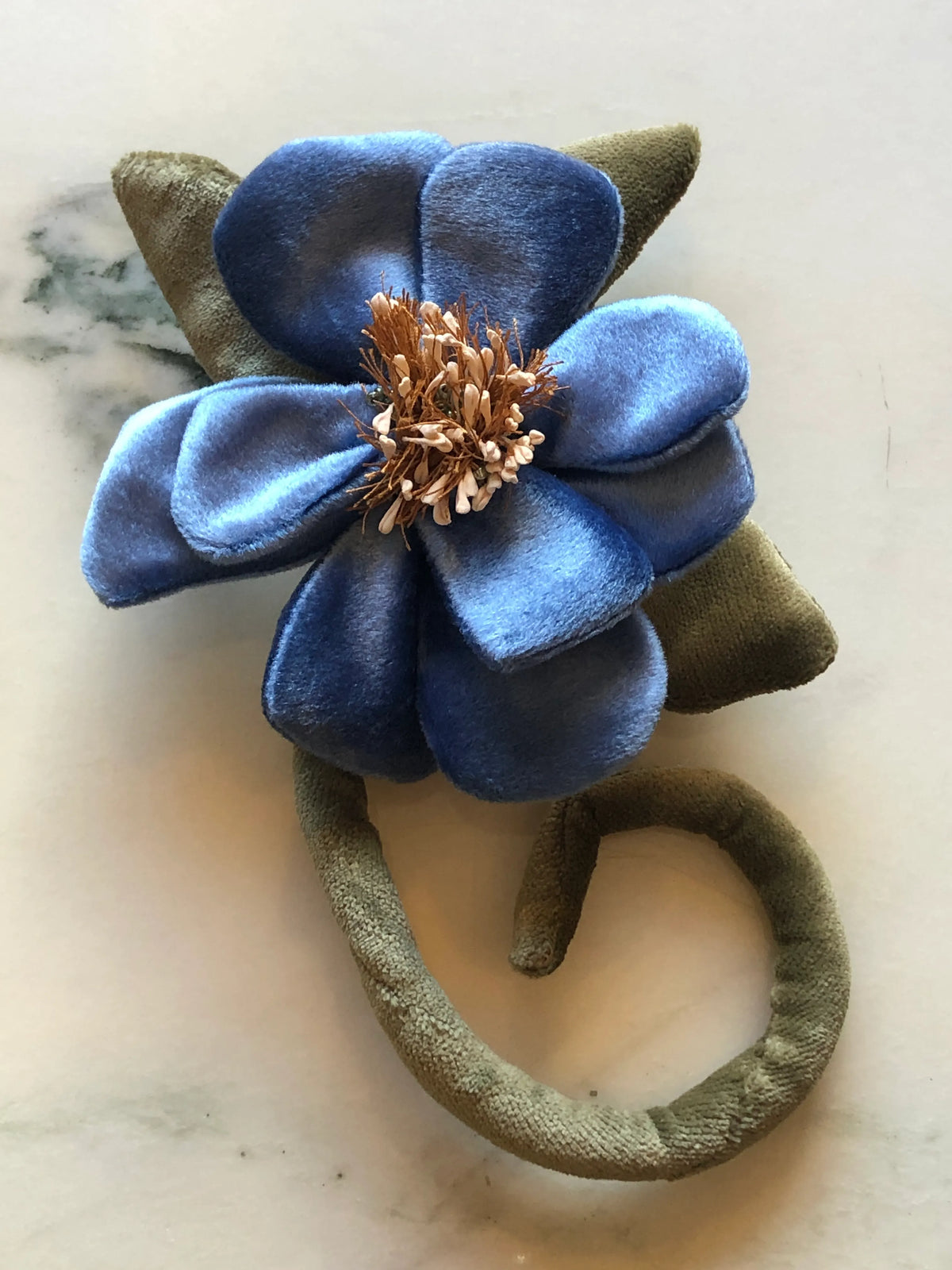 A Broochella Men's Velluto handmade fabric flower brooch with blue velvet petals, olive green velvet leaves and stem, and a metallic bronze center, displayed on a light marble surface.