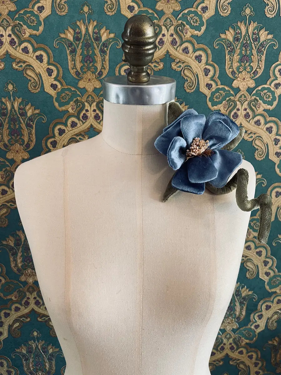 Broochella Velluto blue and  green handmade velvet flower designer brooch with stem and leaves. Styled on a mannequin. The background is green and gold damask wallpaper