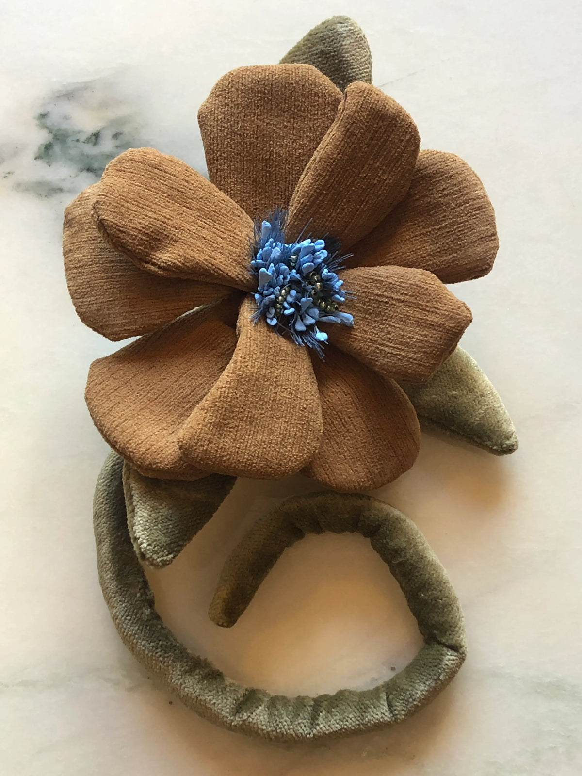 A Broochella handmade velvet fabric flower brooch featuring large brown petals, olive green leaves, and a blue flower centre,  displayed on a light marble surface.