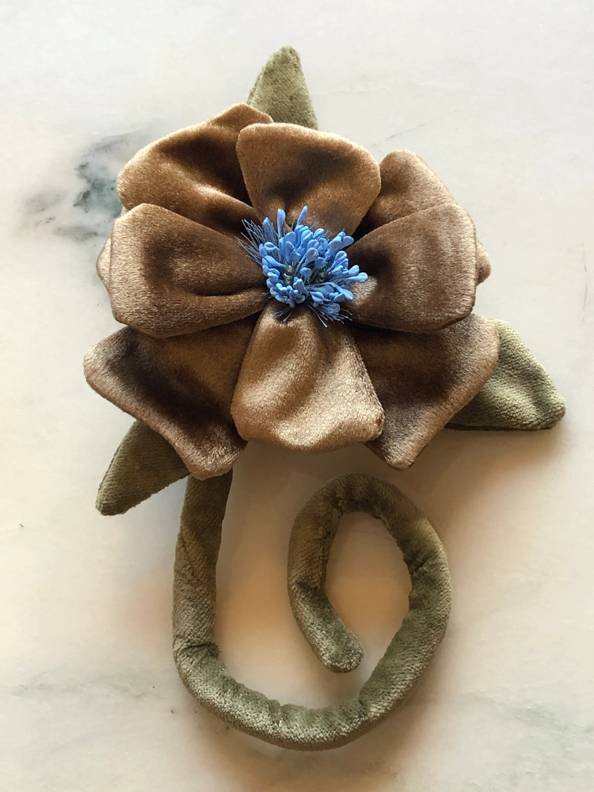 A Broochella Men's Velluto handmade fabric flower brooch featuring a large gold velvet flower with a blue velvet center, olive green leaves, and a matching olive green elastic band, displayed on a light marble surface.