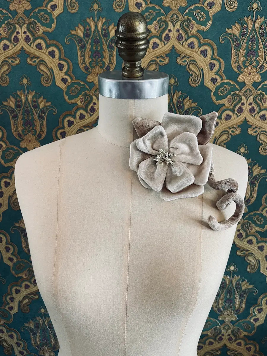 Broochella Velluto ivory and  green handmade velvet flower designer brooch with stem and leaves. Styled on a mannequin. The background is green and gold damask wallpaper