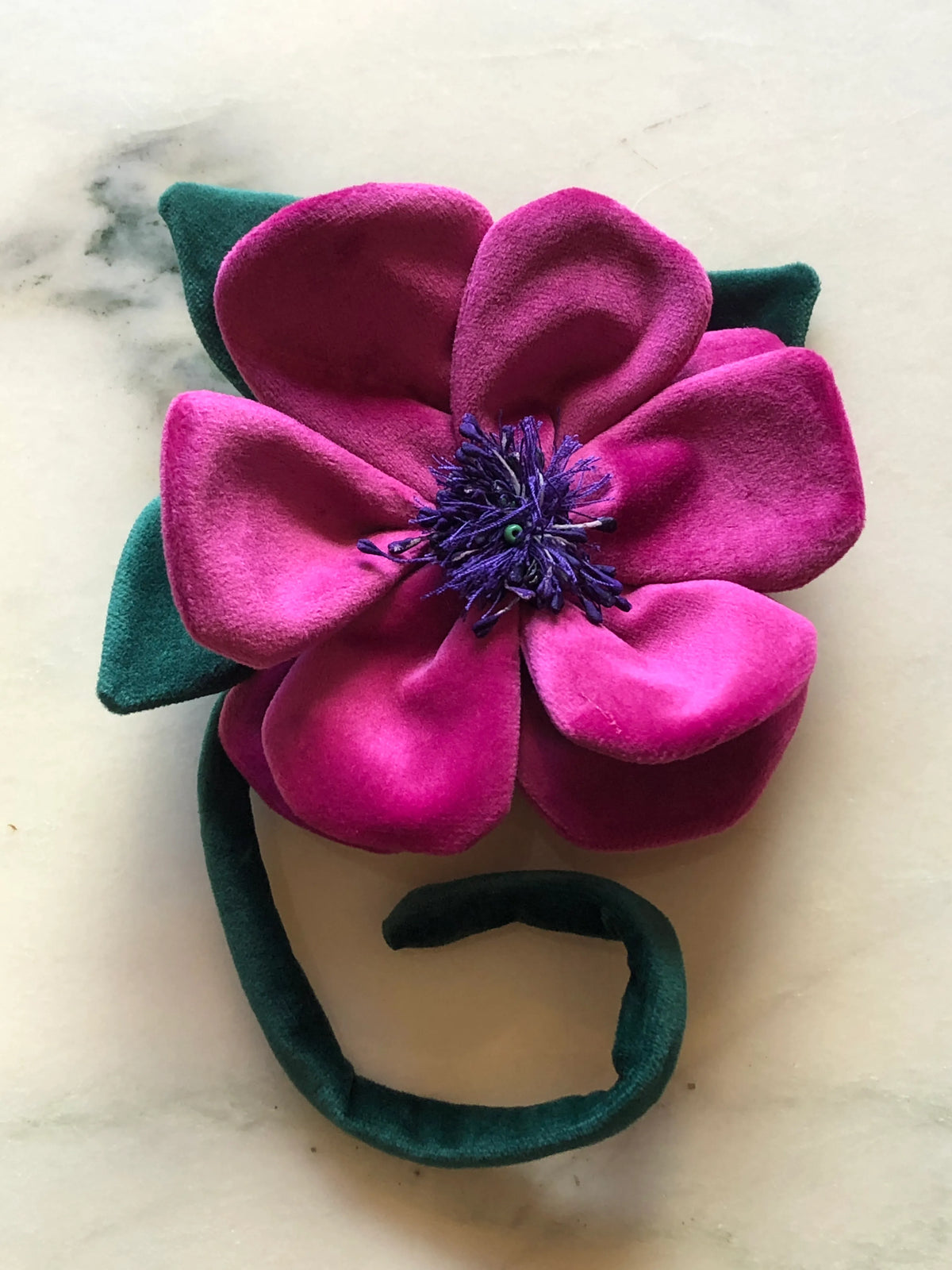 A Broochella Men's Velluto handmade velvet flower brooch featuring a large magenta-pink flower with dark green leaves and a purple centre, attached to a matching dark green velvet headband, displayed on a light marble surface.