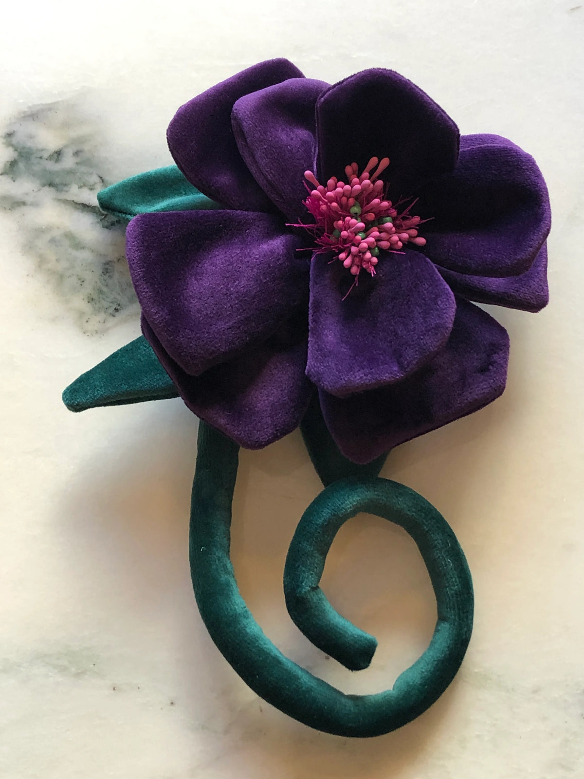 A  Broochella Men's Velluto purple velvet flower brooch with a pink stamen center and a twisted teal velvet stem, displayed on a light marble surface.