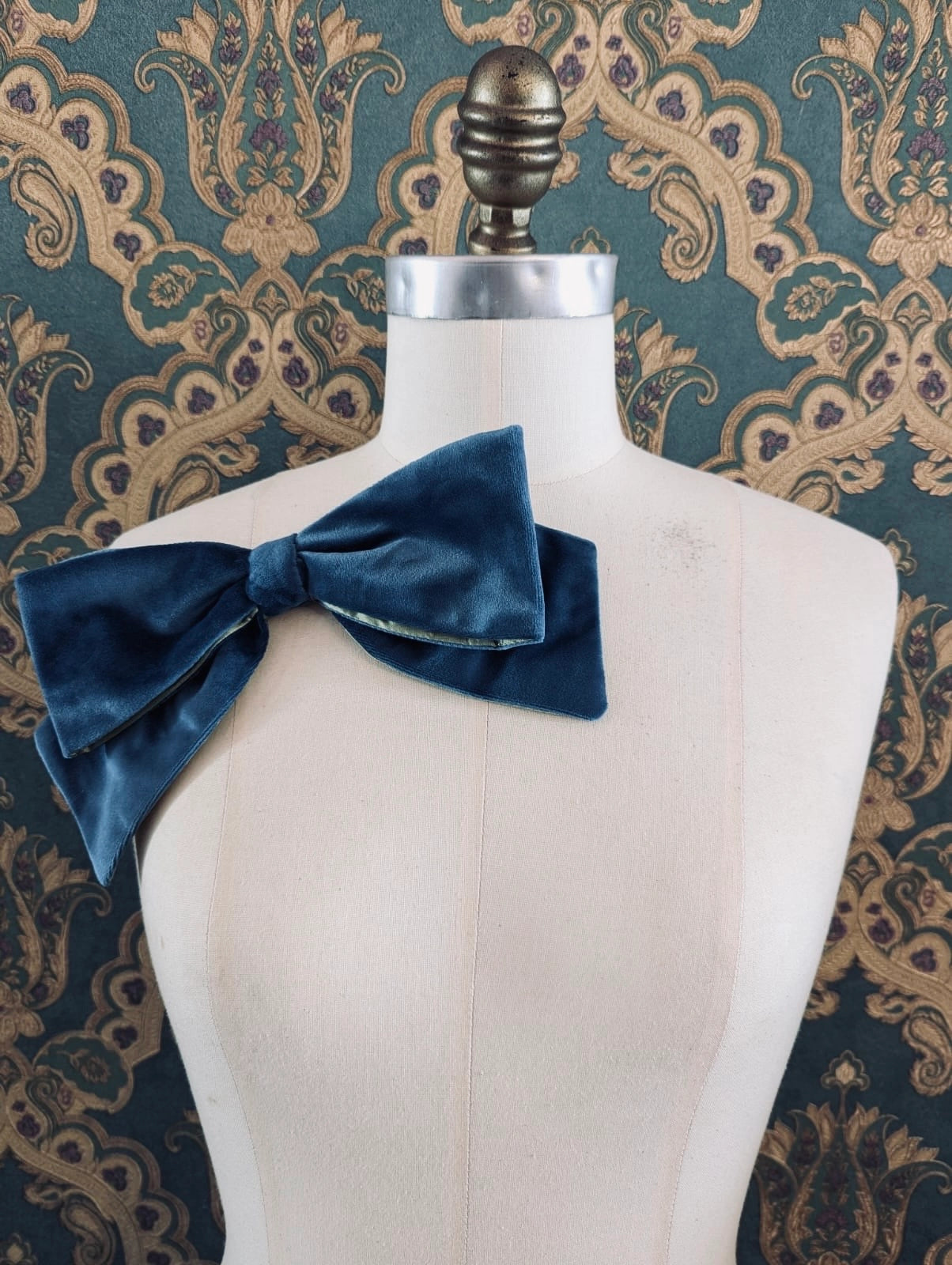 Broochella large velvet handmade designer bow brooch in blue and lime. Pictured on a mannequin.  The background is green and gold damask wallpaper.