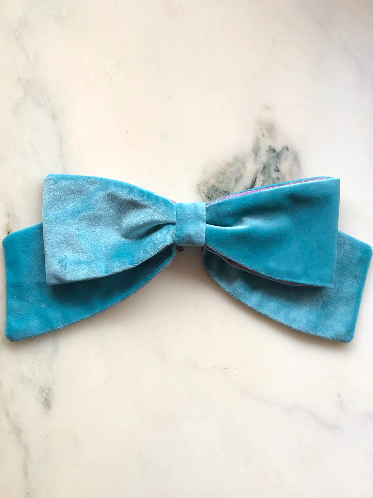 Broochella handmade Large Velvet Bow Brooch in aqua blue velvet with contrasting purple edging, featuring an oversized designer double bow, photographed on white marble.