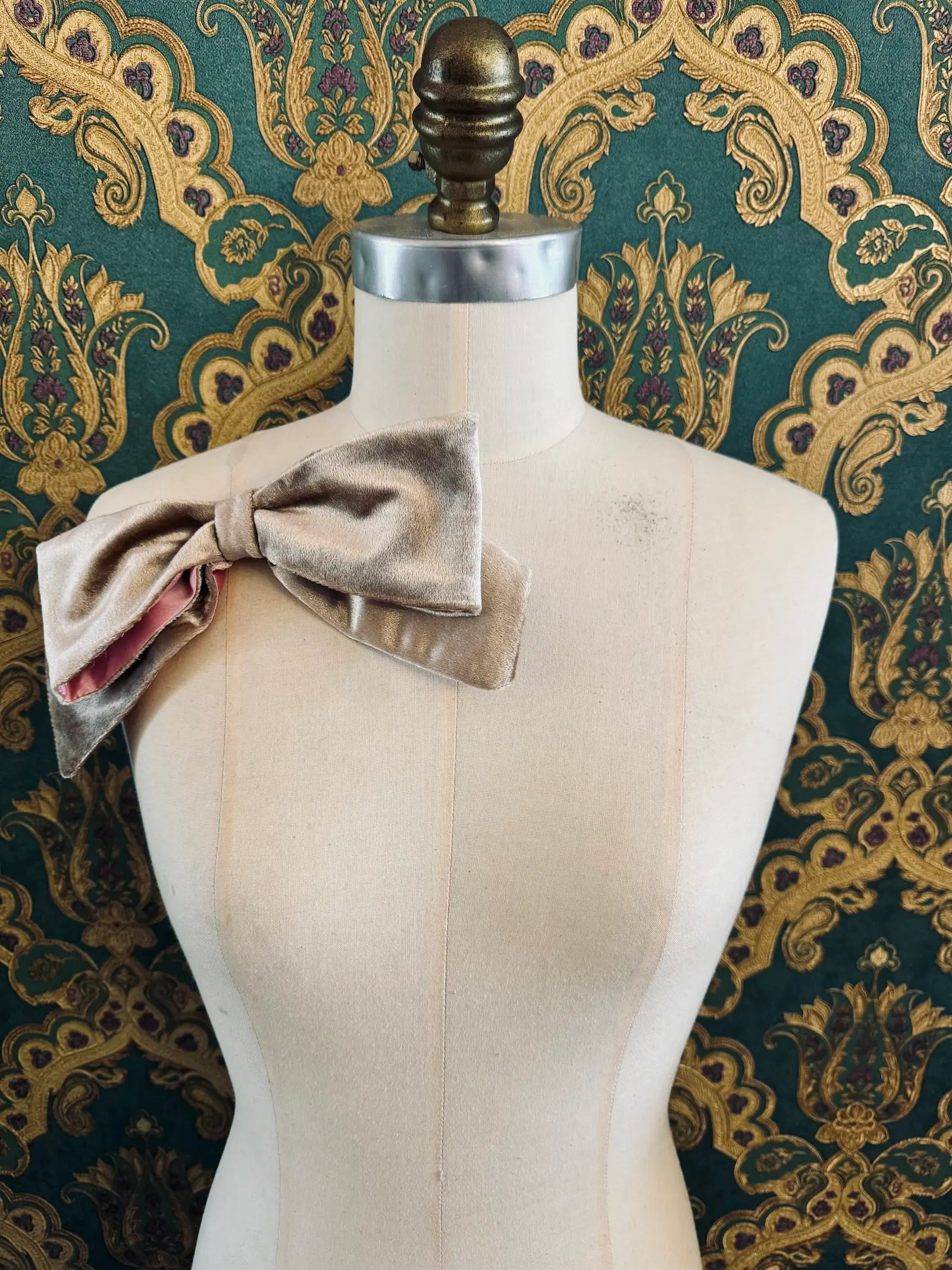 Broochella large velvet handmade bow brooch in champagne and pink. Pictured on a mannequin.  The background is green and gold damask wallpaper.