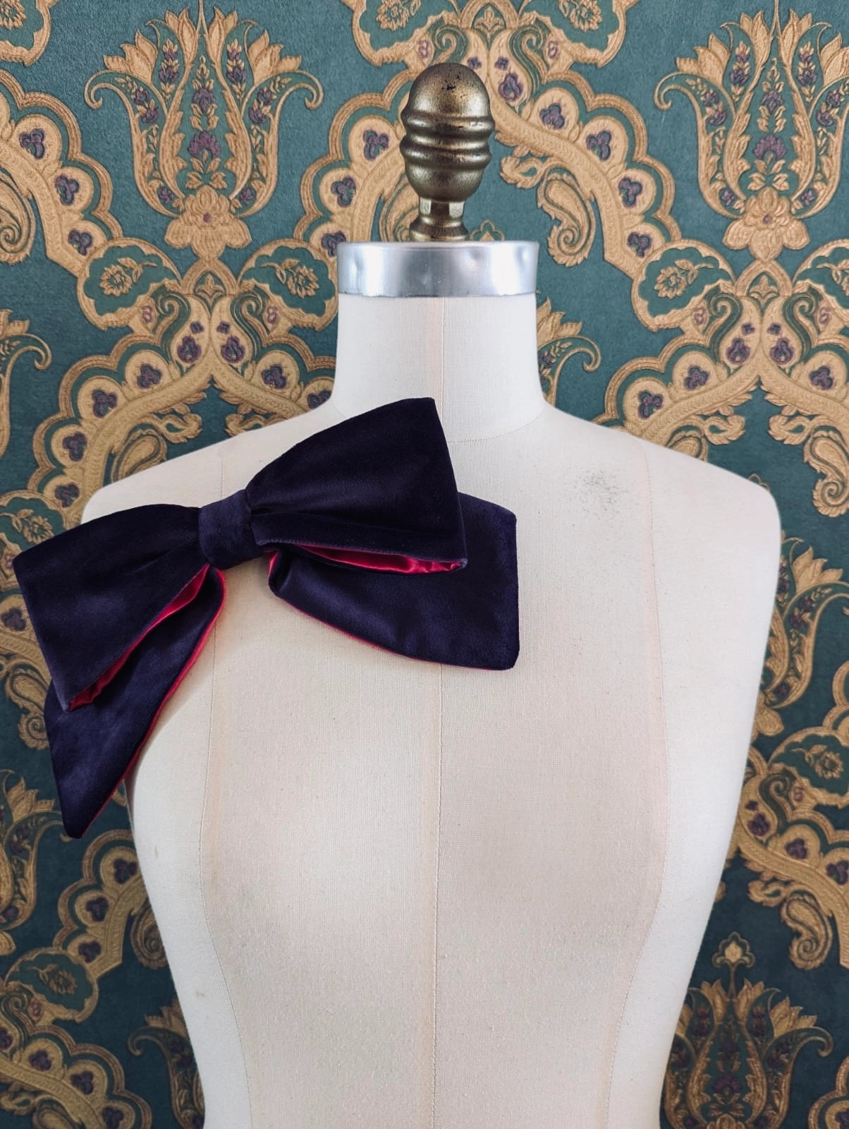 Broochella large velvet handmade designer bow brooch in purple and fuchsia. Pictured on a mannequin.  The background is green and gold damask wallpaper.