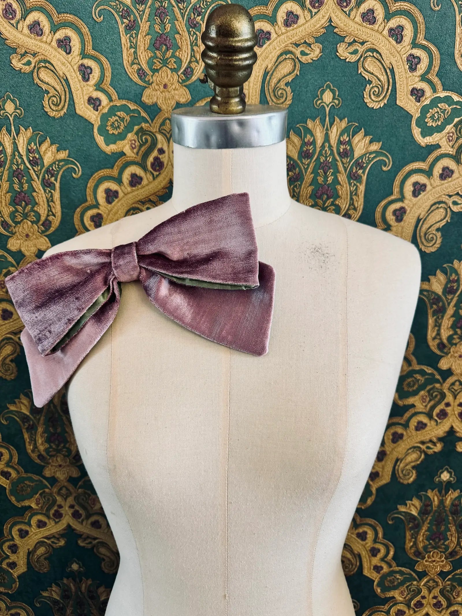 Broochella large velvet handmade bow brooch in violet and green. Pictured on a mannequin.  The background is green and gold damask wallpaper.