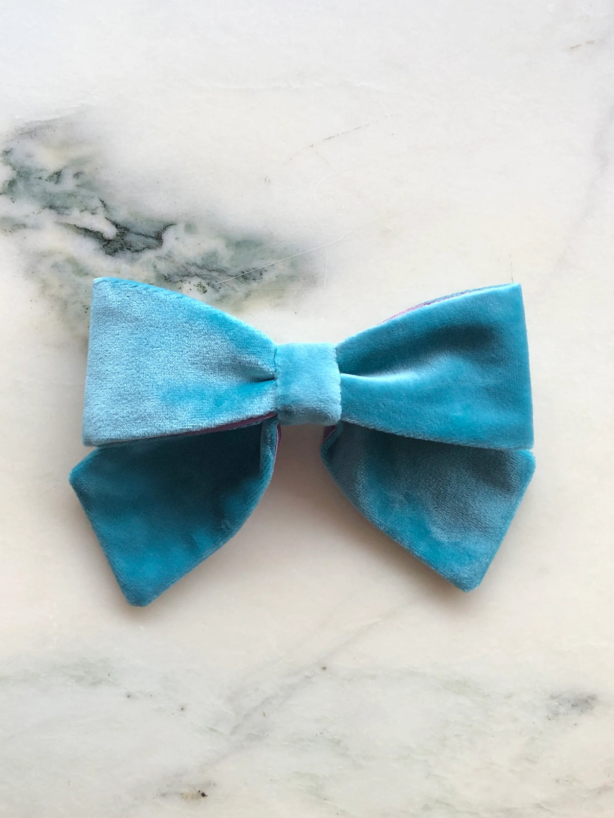 Broochella men's handmade Small Velvet Bow Brooch in aqua blue velvet with contrasting purple edging, featuring a designer double bow, photographed on white marble.