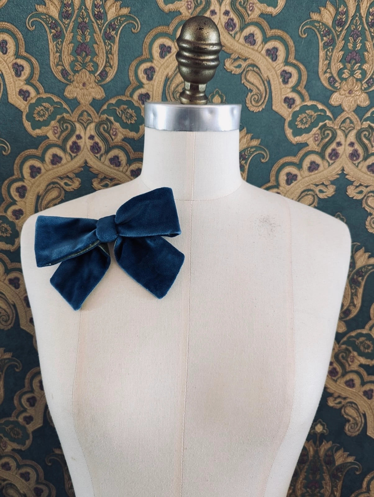 Broochella small velvet handmade designer bow brooch in blue and lime. Pictured on a mannequin.  The background is green and gold damask wallpaper.
