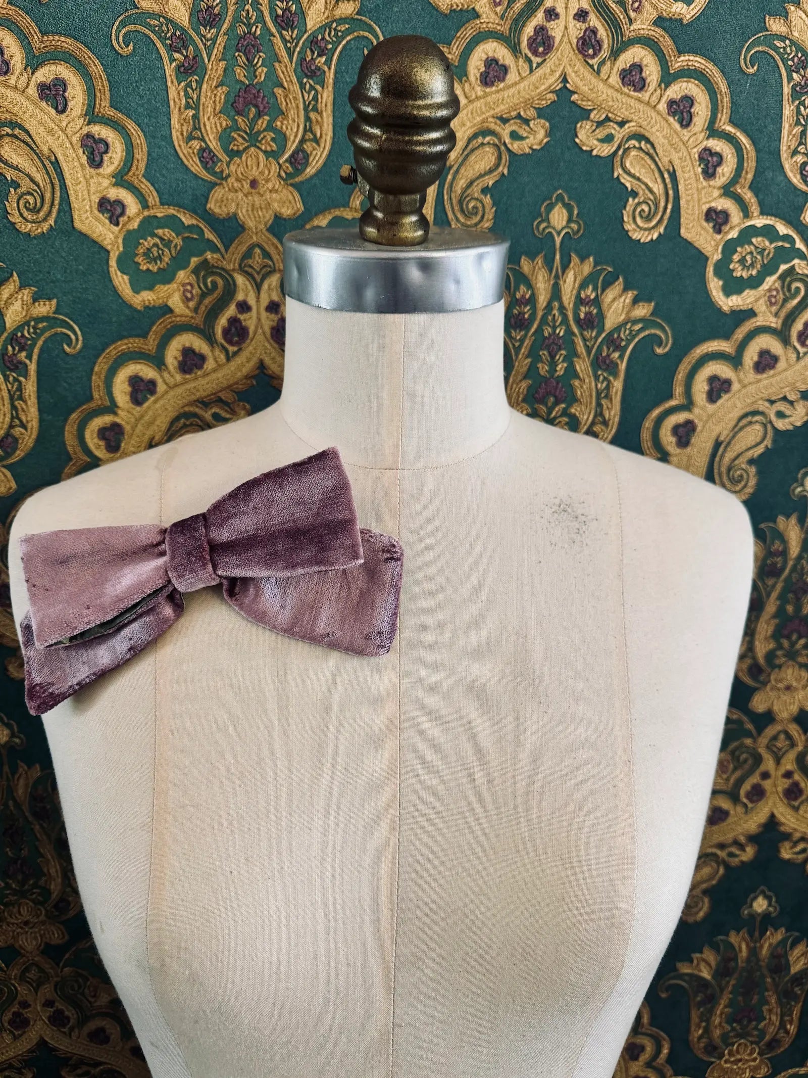 Broochella small velvet handmade bow brooch in violet and green. Pictured on a mannequin.  The background is green and gold damask wallpaper.