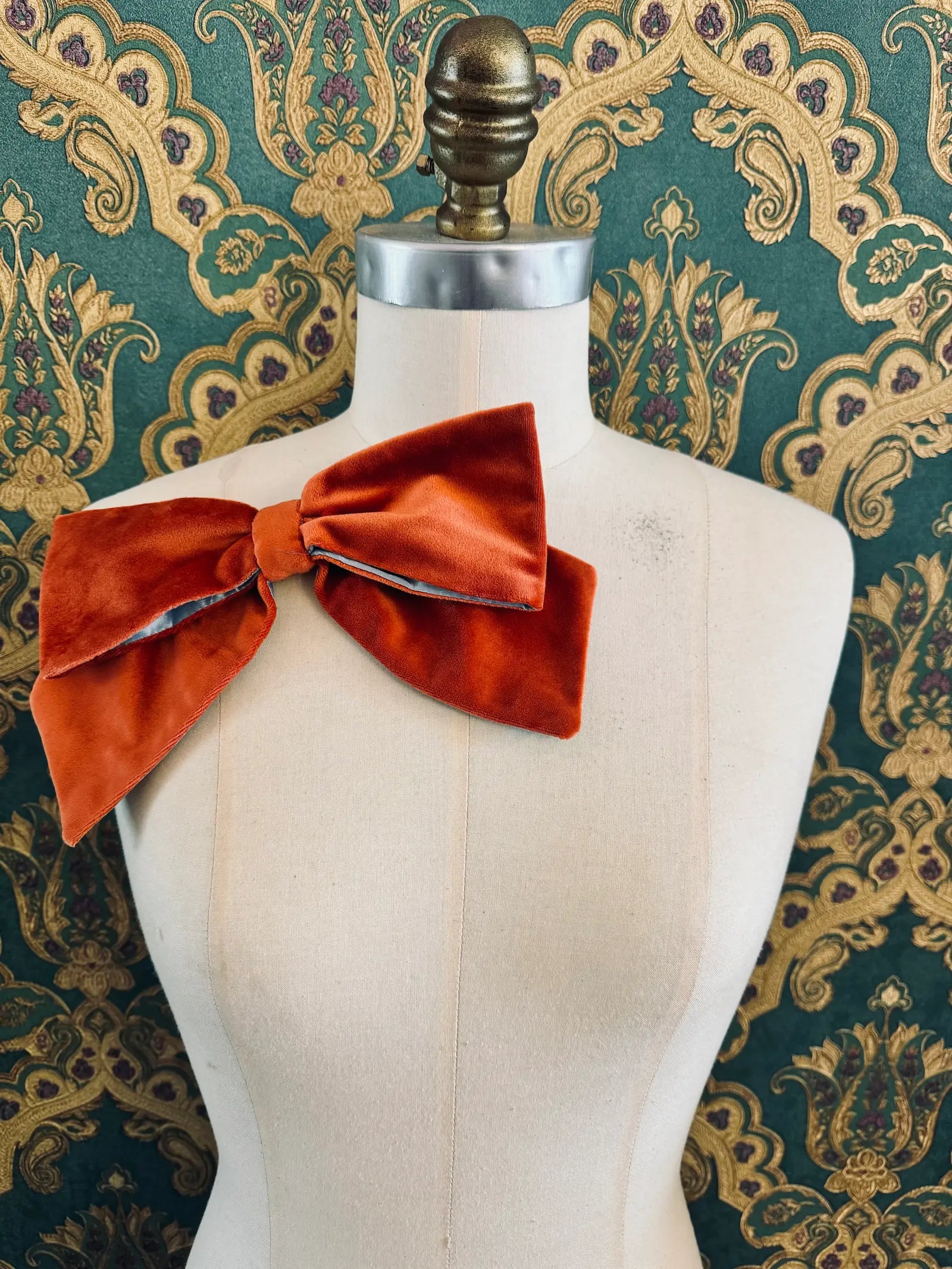 Broochella large velvet handmade bow brooch in orange and ice blue. Pictured on a mannequin.  The background is green and gold damask wallpaper.