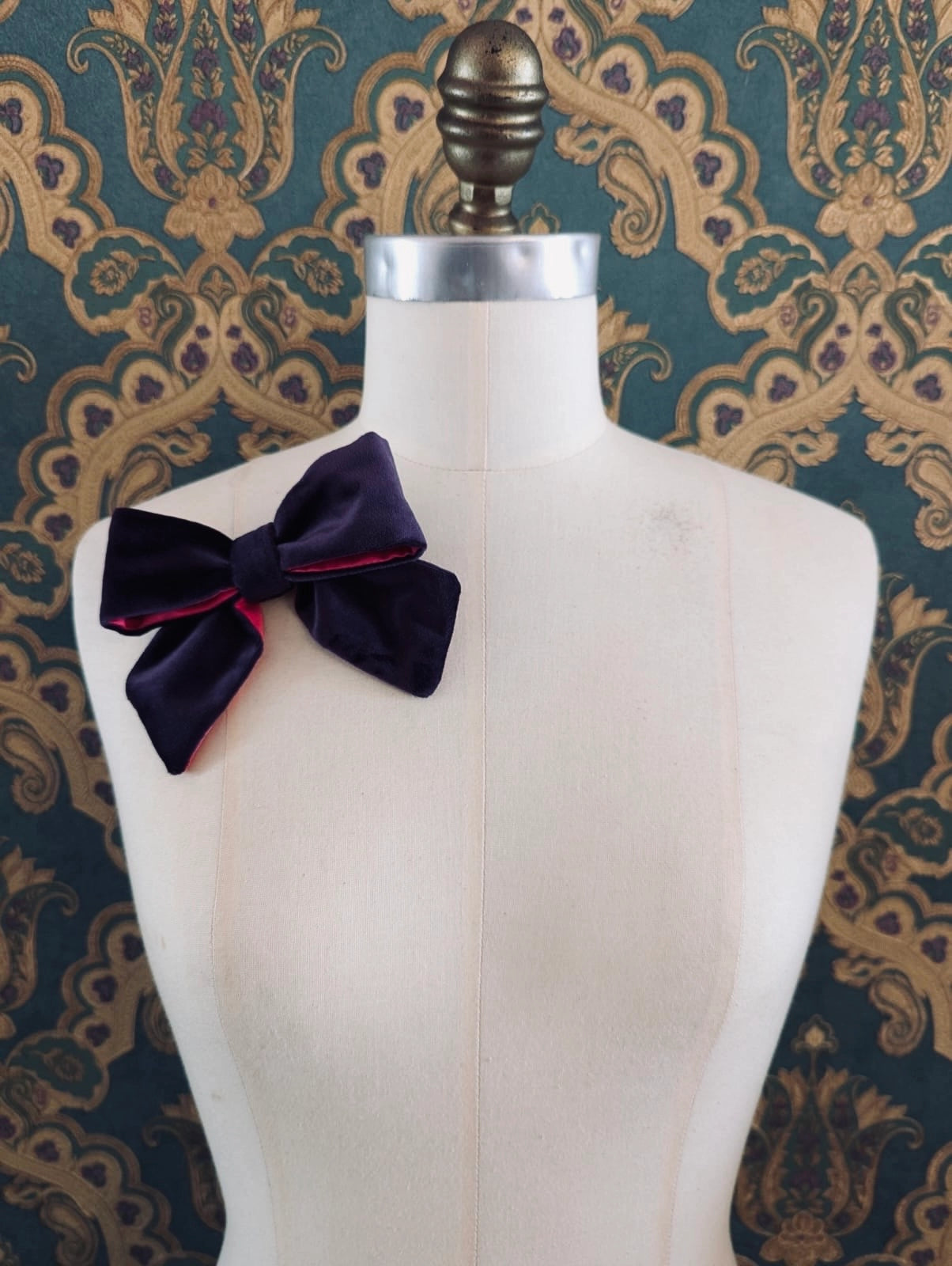 Broochella small velvet handmade designer bow brooch in purple and fuchsia. Pictured on a mannequin.  The background is green and gold damask wallpaper.
