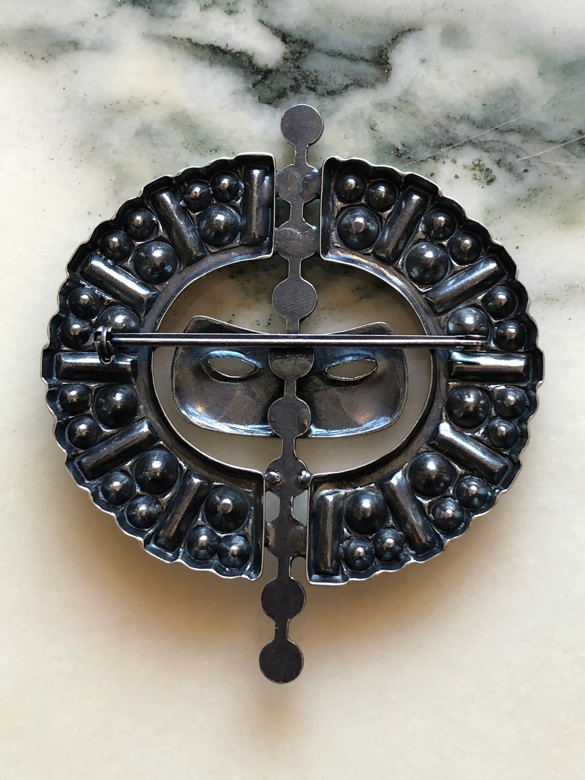 Broochella Venezia Vintage Masquerade Crystal Brooch featuring a silver-tone mask surrounded by blue, red and green crystals in an ornate circular setting on a cream and grey marble background.