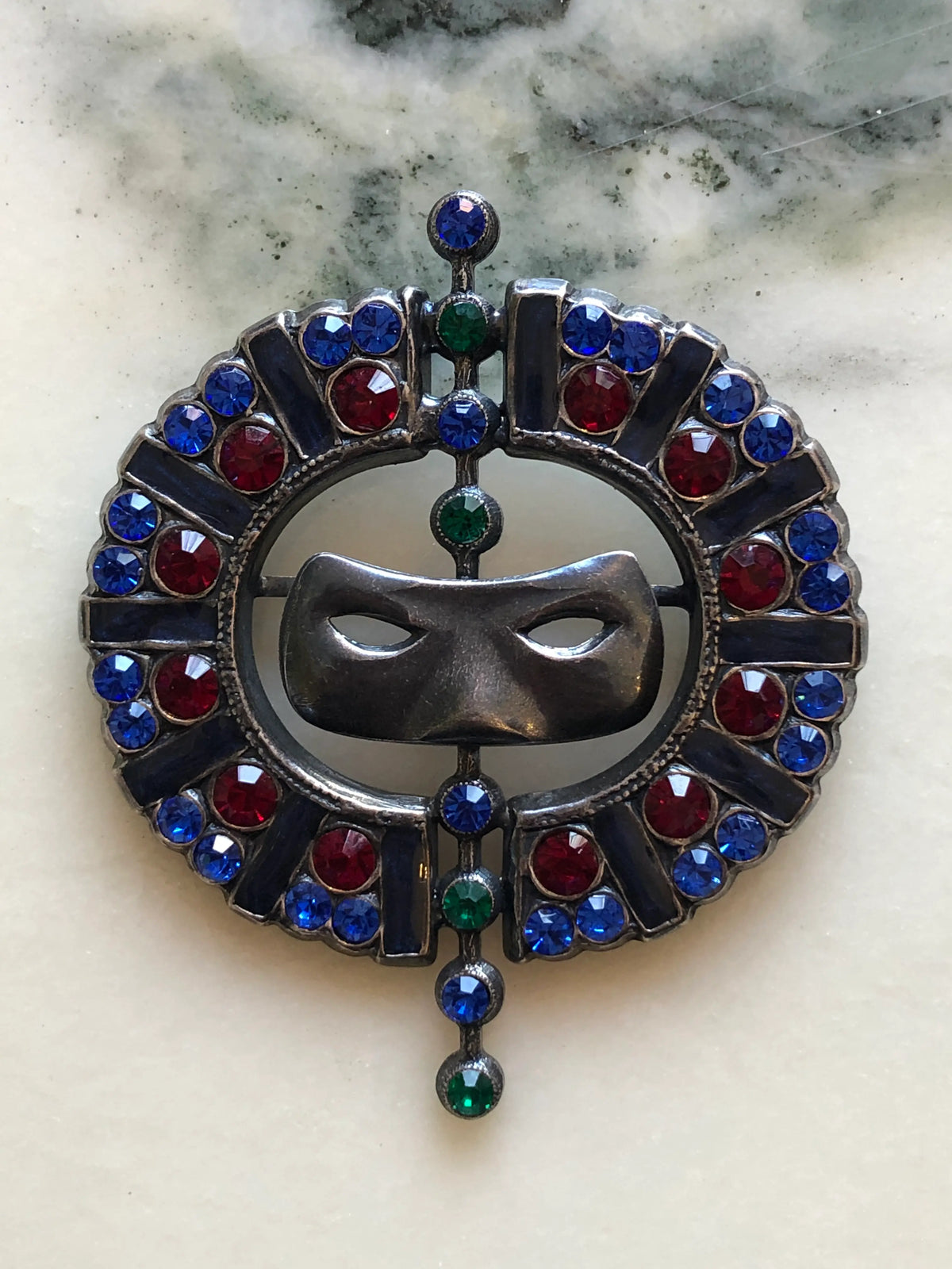 Broochella Venezia Vintage Masquerade Crystal Brooch featuring a silver-tone mask surrounded by blue, red and green crystals in an ornate circular setting on a cream and grey marble background.