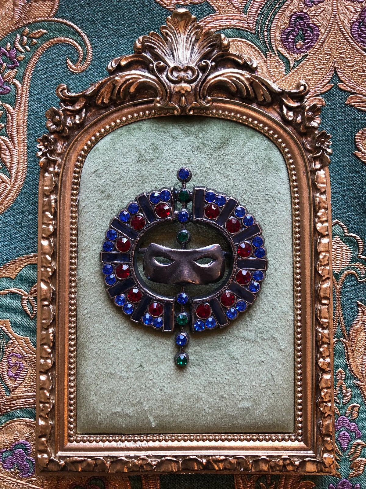 Broochella Venezia Vintage Masquerade Crystal Brooch featuring a silver-tone mask surrounded by blue, red and green crystals in an ornate circular setting.  It is presented on a green velvet tray with an ornate gold frame. The background is green and gold damask wallpaper.
