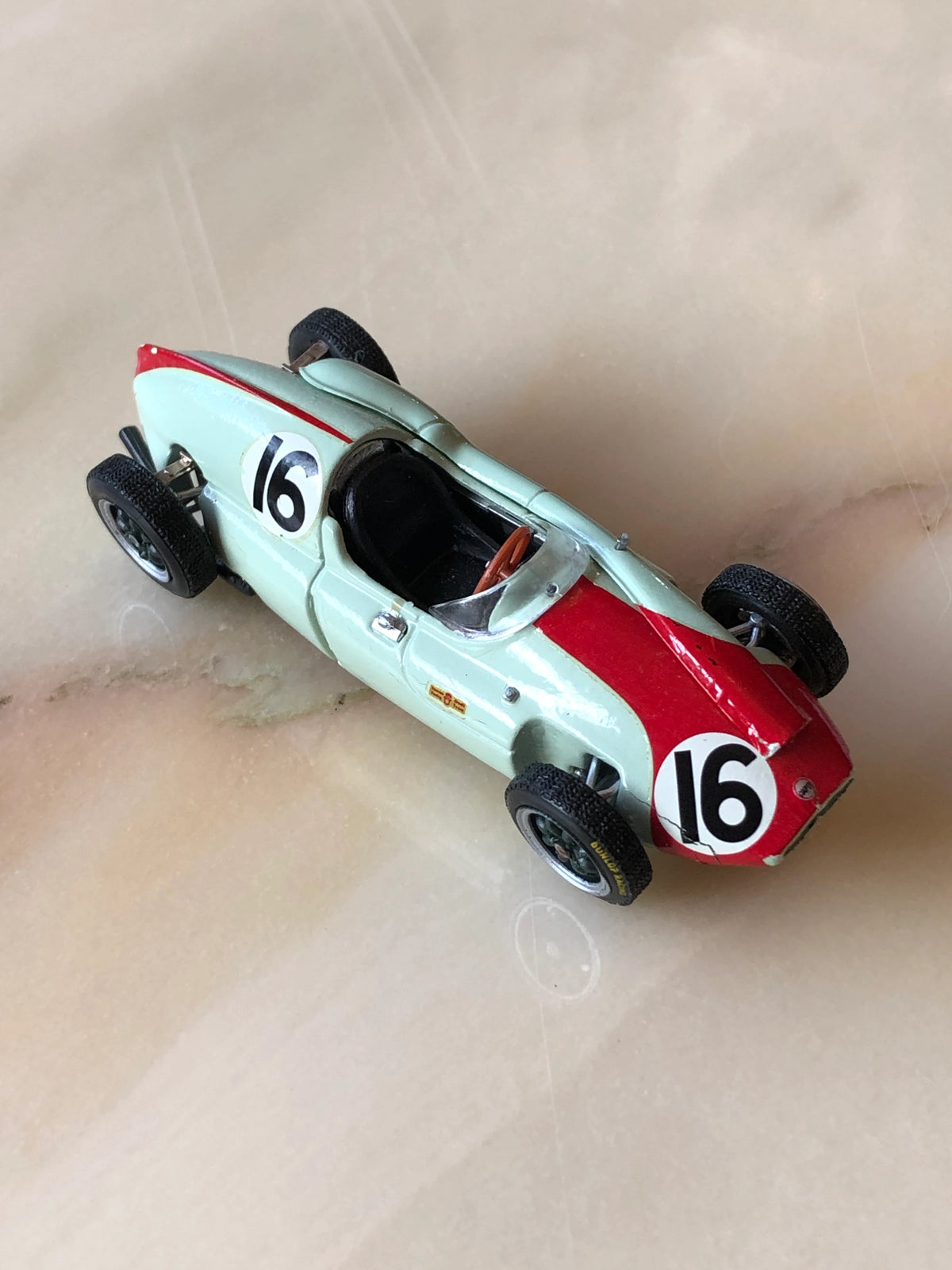 Broochella Verde Corsa No.16 Vintage Formula Racing Car Brooch photographed at an elevated angle, showcasing its mint green body, bold red bonnet stripe, number 16 racing decals, brown steering wheel and detailed black tyres.