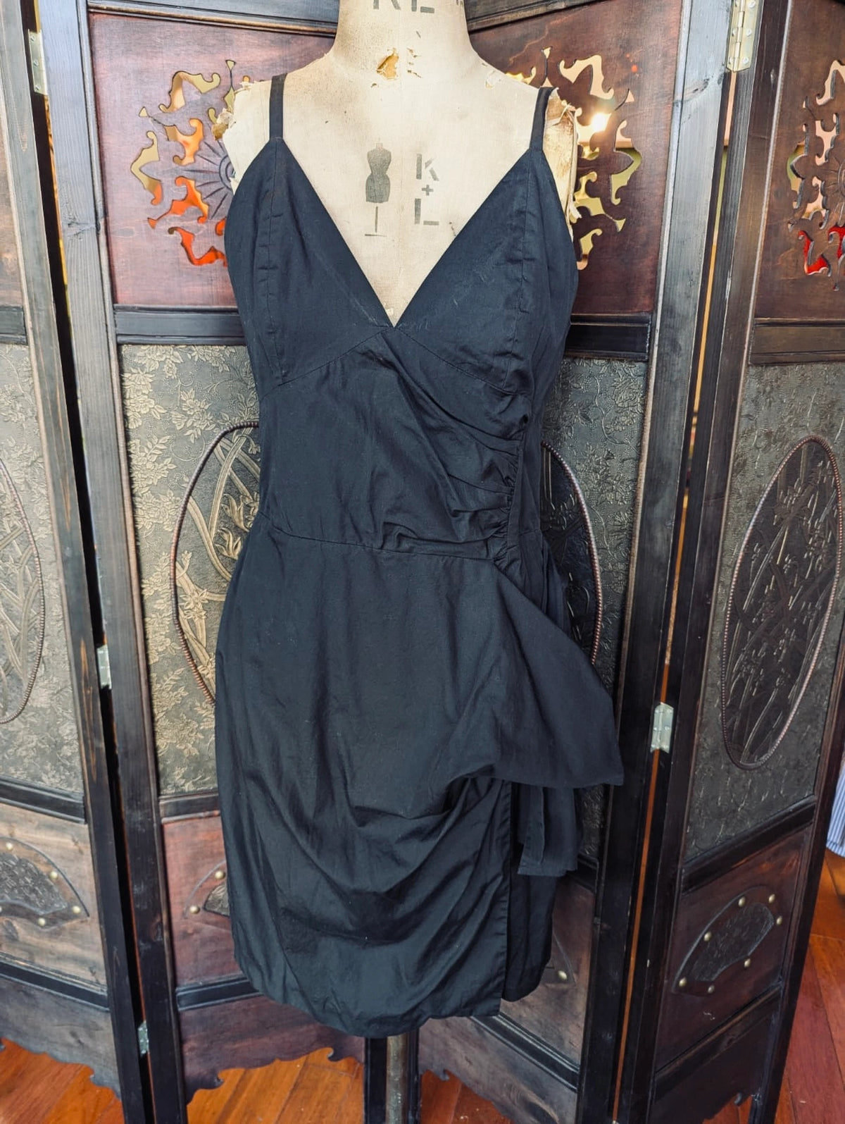 A Broochella Victoria Beckham black sleeveless wrap-style dress with thin straps and a gathered peplum-like hem, displayed on a mannequin torso in front of a decorative wooden folding screen.