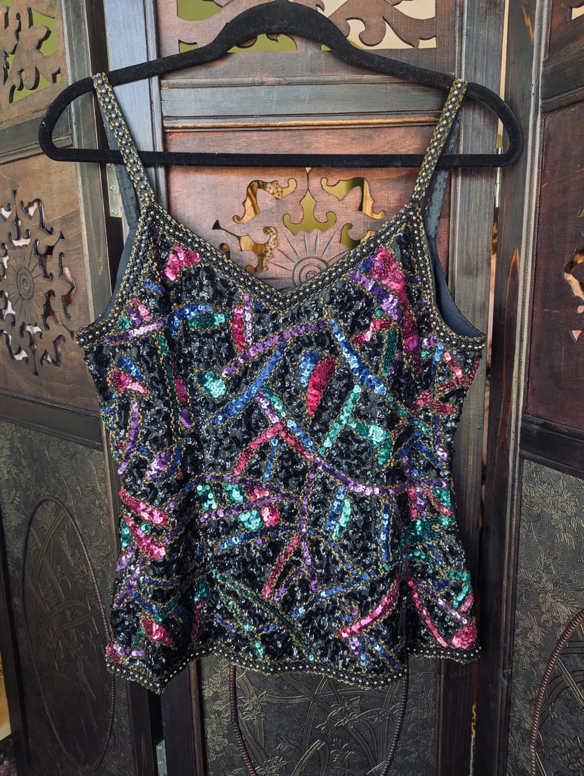 A Broochella Vintage 1980's black sequinned camisole top with multicoloured iridescent sequins and gold-tone trim, hanging on a black hanger against a dark carved wooden door.