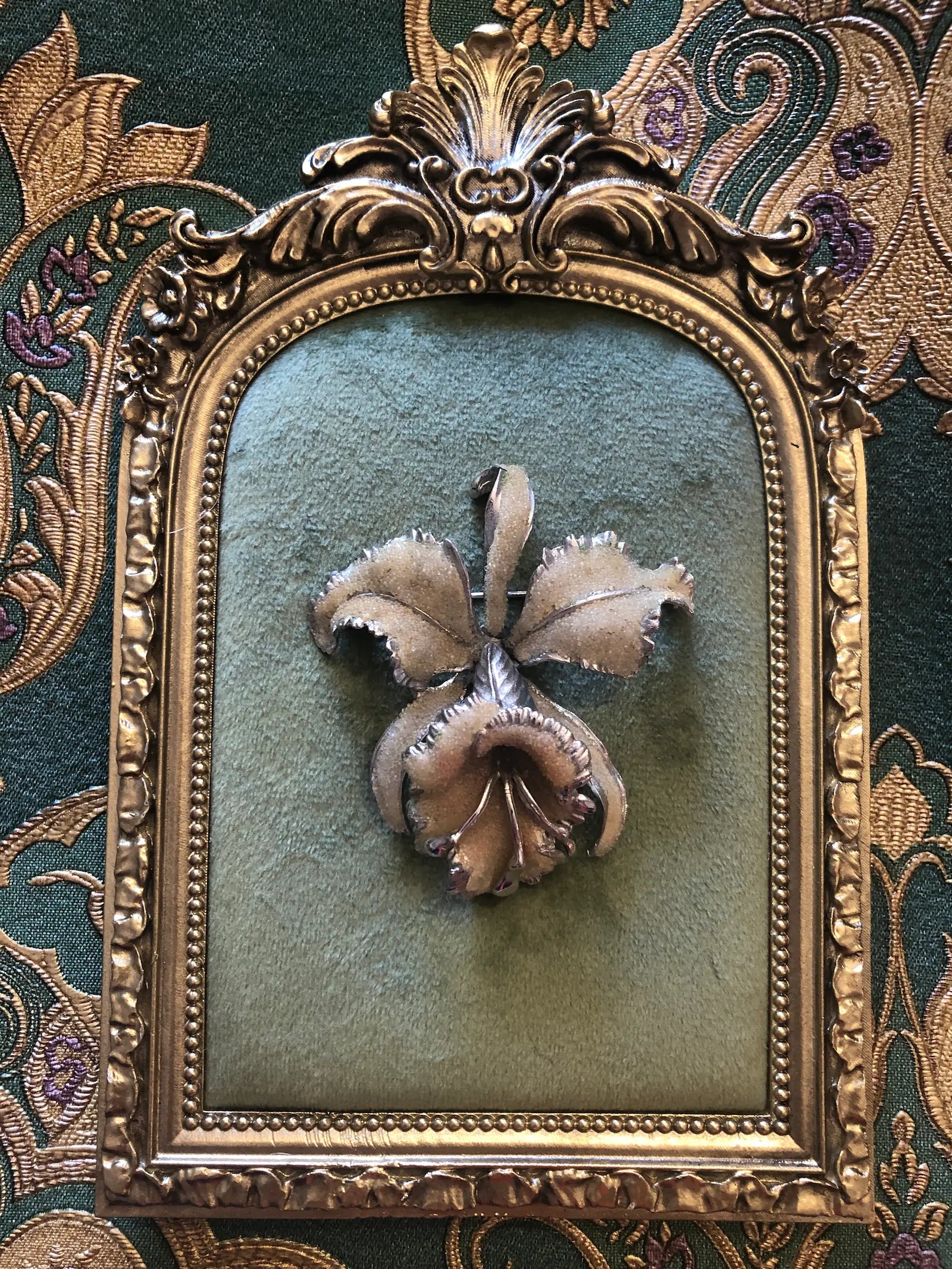 A vintage signed Elizabeth Reimer Arcansas Coralene silver-tone orchid brooch featuring five sculptural petals with a fine frosted sand-texture coralene surface, polished serrated silver-tone metal petal edges and a three-dimensional trumpet centre, photographed on a gold frame with green velvet centre. The background is green and gold damask wallpaper.