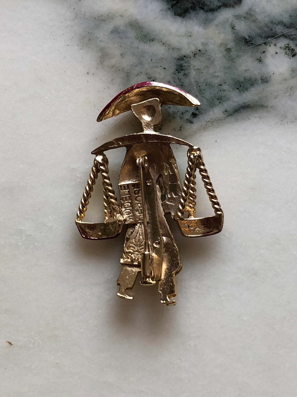 Broochella Vintage Enamel Water Carrier Brooch - Green featuring a figural water carrier in vivid green enamel with a raspberry-red hat, suspended vessels and gold-tone detailing on a cream and grey marble background.
