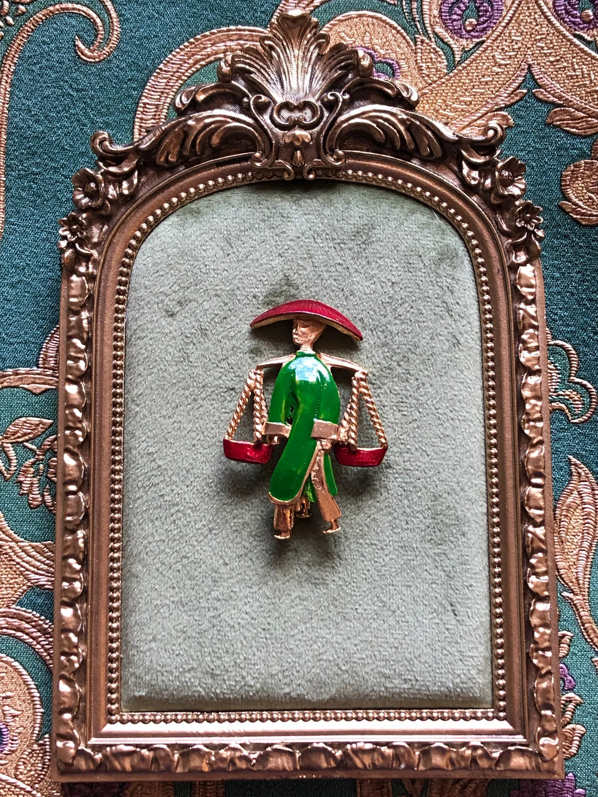 Broochella Vintage Enamel Water Carrier Brooch - Green featuring a figural water carrier in vivid green enamel with a raspberry-red hat, suspended vessels and gold-tone detailing on a cream and grey marble background.