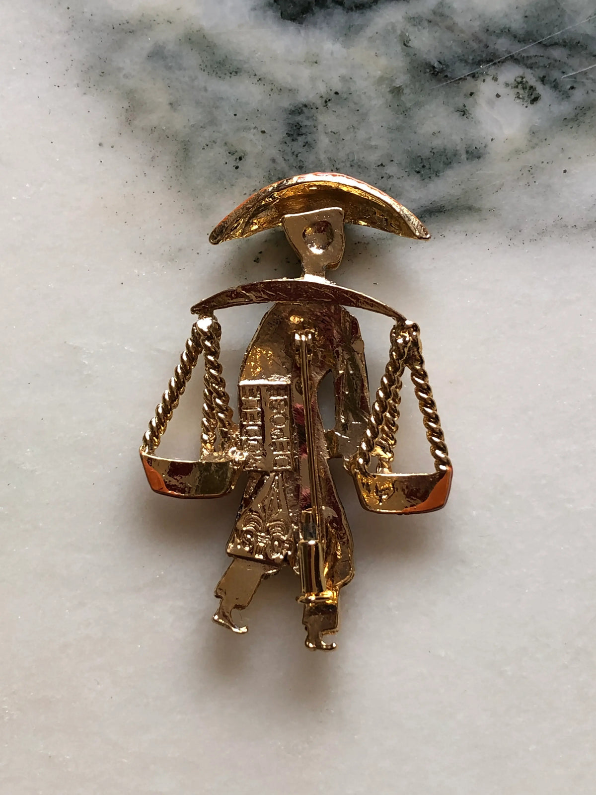 The back view Broochella Vintage Enamel Water Carrier Brooch - Navy featuring a figural water carrier in deep navy enamel with an orange hat, suspended vessels and gold-tone detailing on a cream and grey marble background.