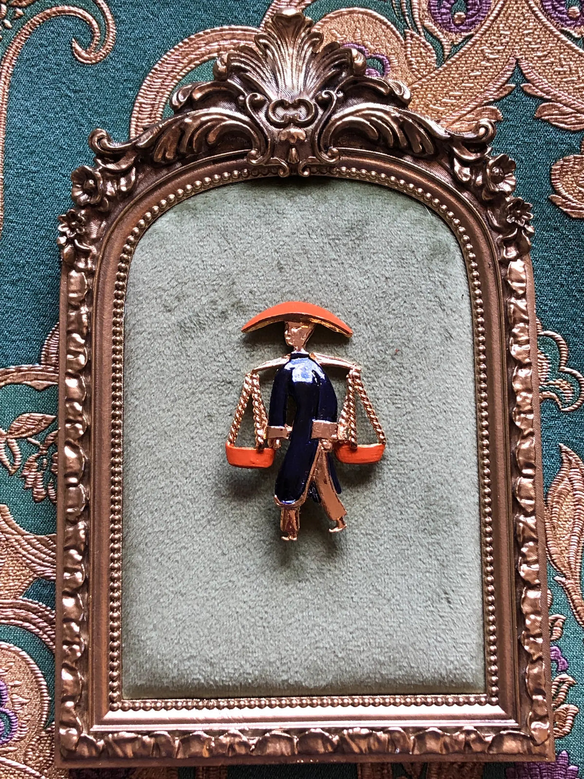 Broochella Vintage Enamel Water Carrier Brooch - Navy featuring a figural water carrier in deep navy enamel with an orange hat, suspended vessels and gold-tone detailing. Pictured on green velvet tray with an ornate gold frame. The background is green and gold damask wallpaper