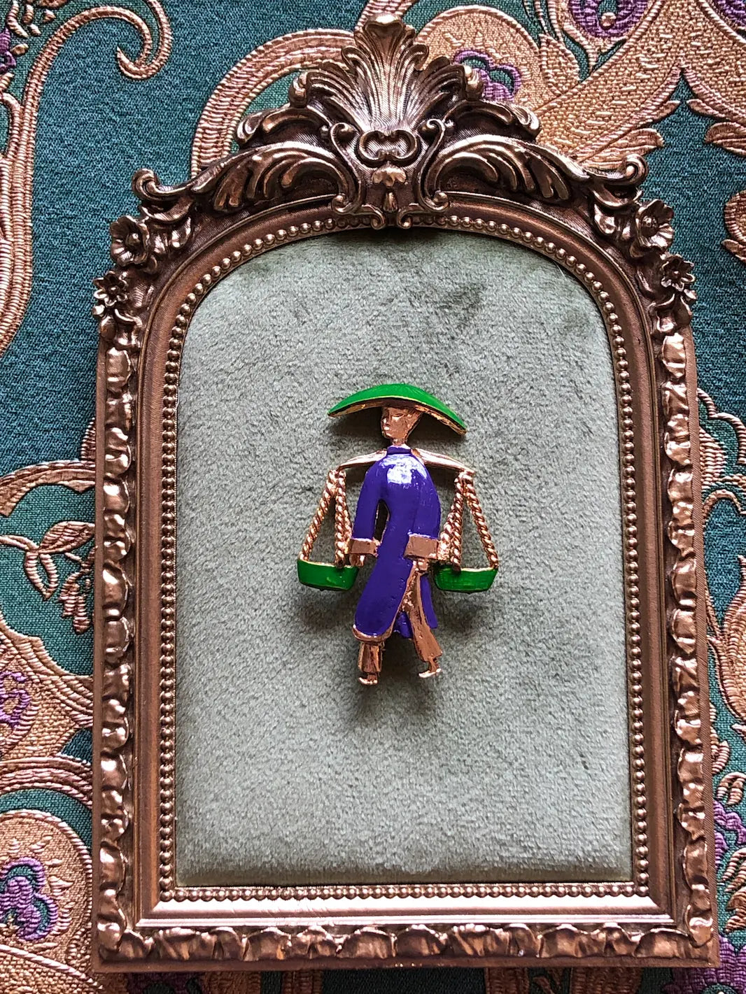Broochella Vintage Enamel Water Carrier Brooch - Purple featuring a figural water carrier in violet-purple enamel with a green hat, suspended green vessels and gold-tone detailing. Pictured on a green velvet tray with an ornate gold frame. The background is green and gold damask wallpaper. 