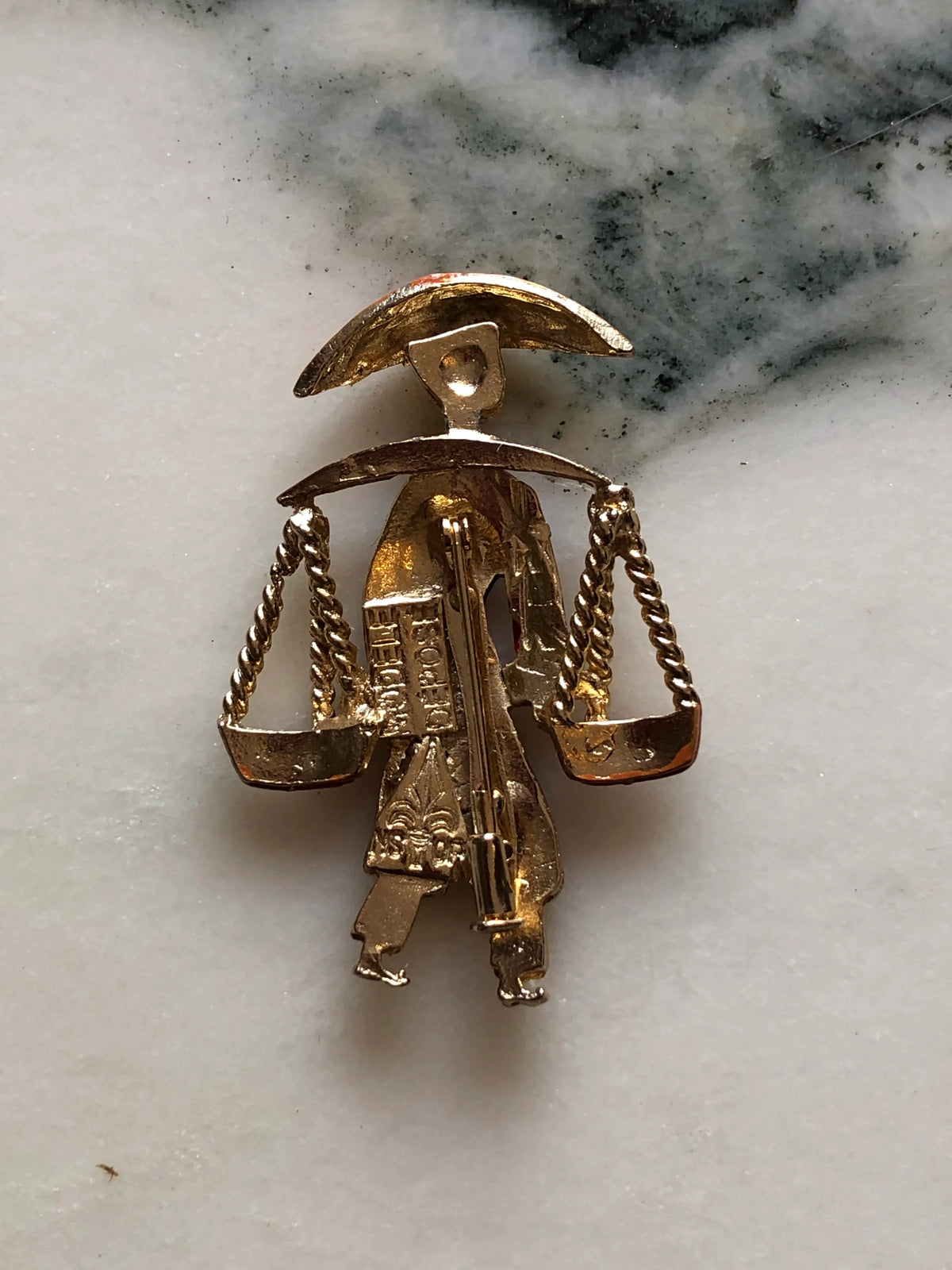 The back view of Broochella Vintage Enamel Water Carrier Brooch featuring a figural water carrier in raspberry-red and orange enamel with suspended vessels and gold-tone detailing on a cream and grey marble background.