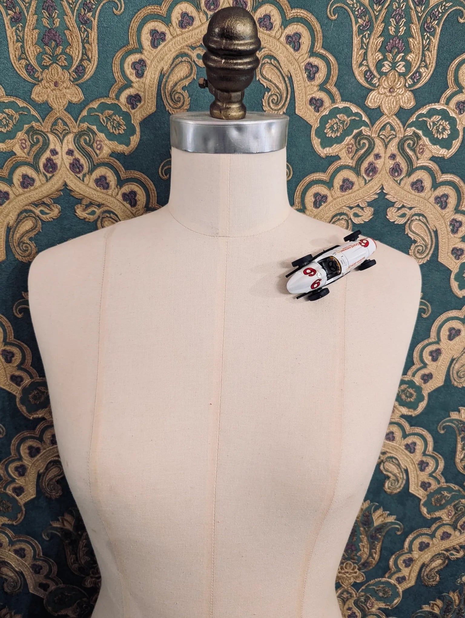 Broochella  scuderia d'oro  bianca numner six vintage formula one racing car brooch. Pinned on a mannequin. The background is green and gold damask wallpaper. 