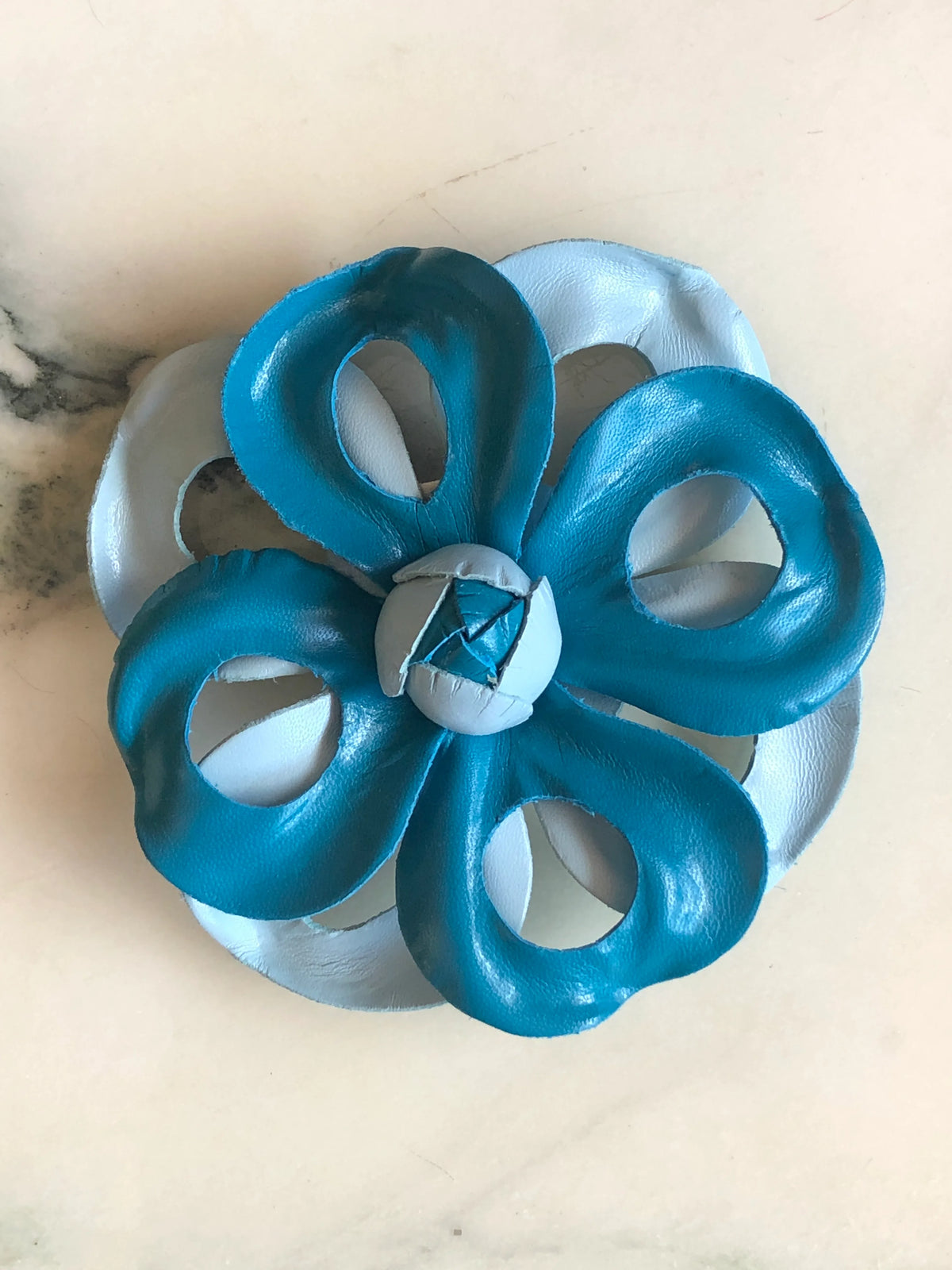 Broochella Viola del Pensiero Leather Flower Brooch Pin in blue, featuring sculptural two-tone turquoise and pale blue leather petals with open cut-out detailing and a handcrafted centre on a cream marble background.